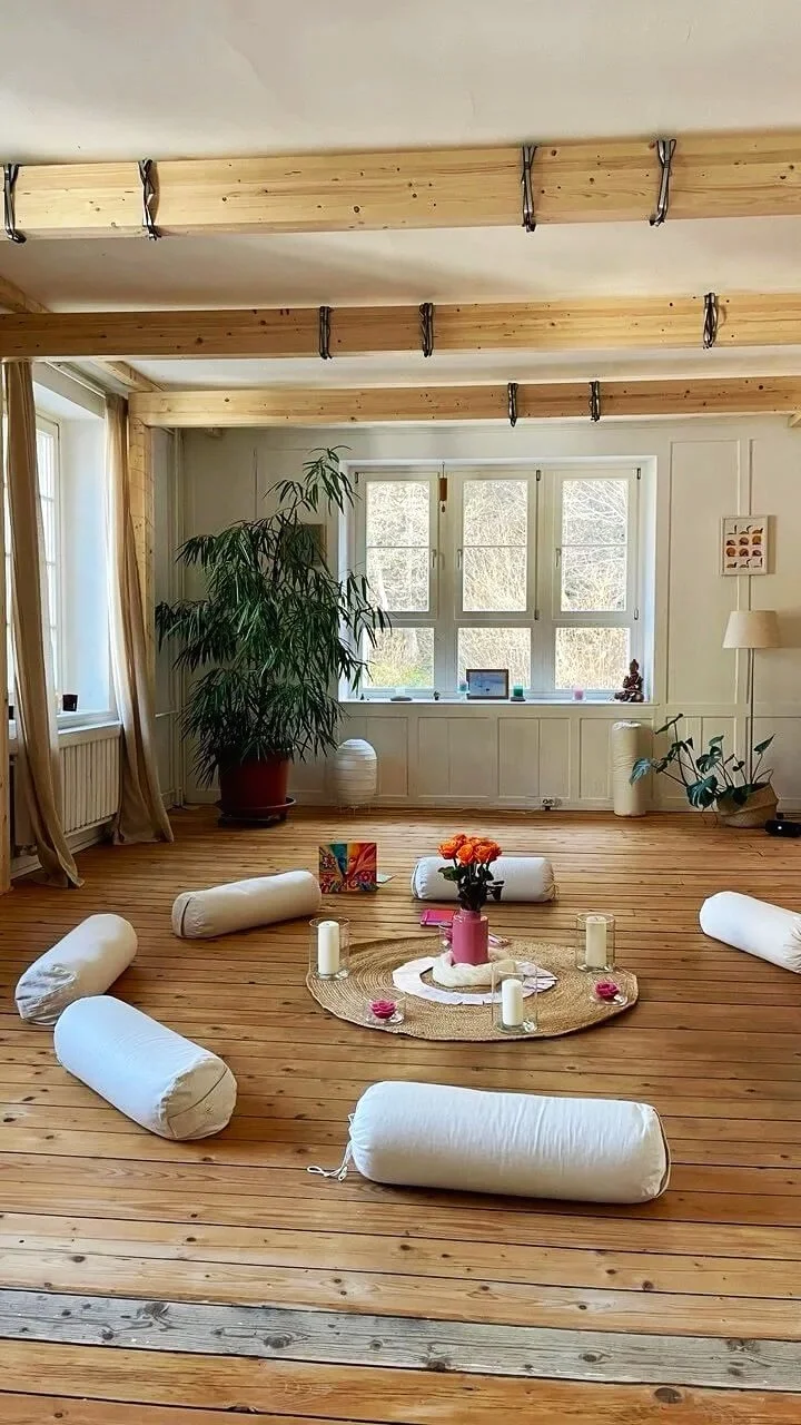 Ein gemütlich eingerichteter Raum mit Holzfußboden, großen Fenstern, einer Pflanzen und meditativen Gegenständen wie Kerzen und Kissen.