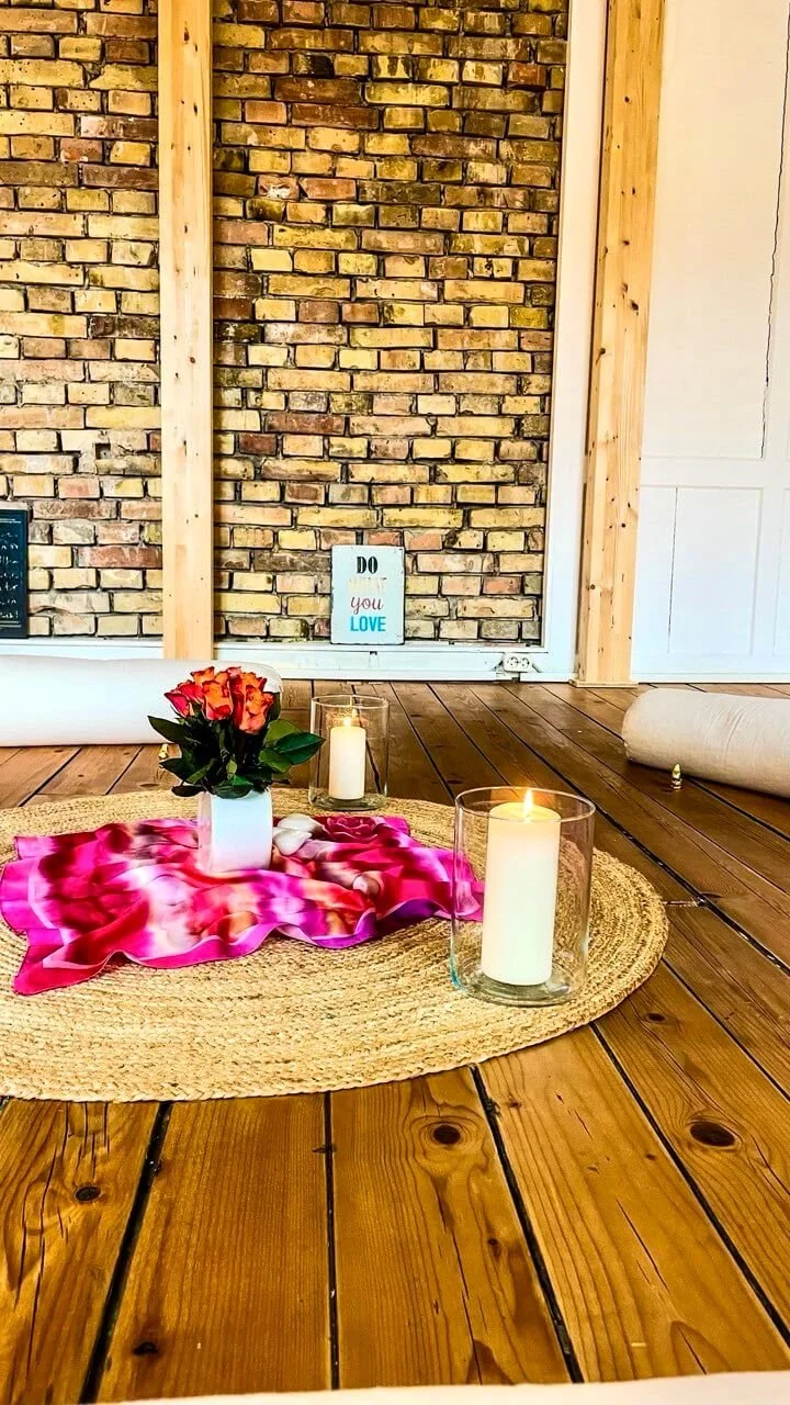 Votivkerzen, Blumenstrauß in weißer Vase, rotes und pinkes Tuch auf einem runden, geflochtenen Teppich vor einer Backsteinwand mit einem kleinen Schild, auf dem 'Do you love' steht.