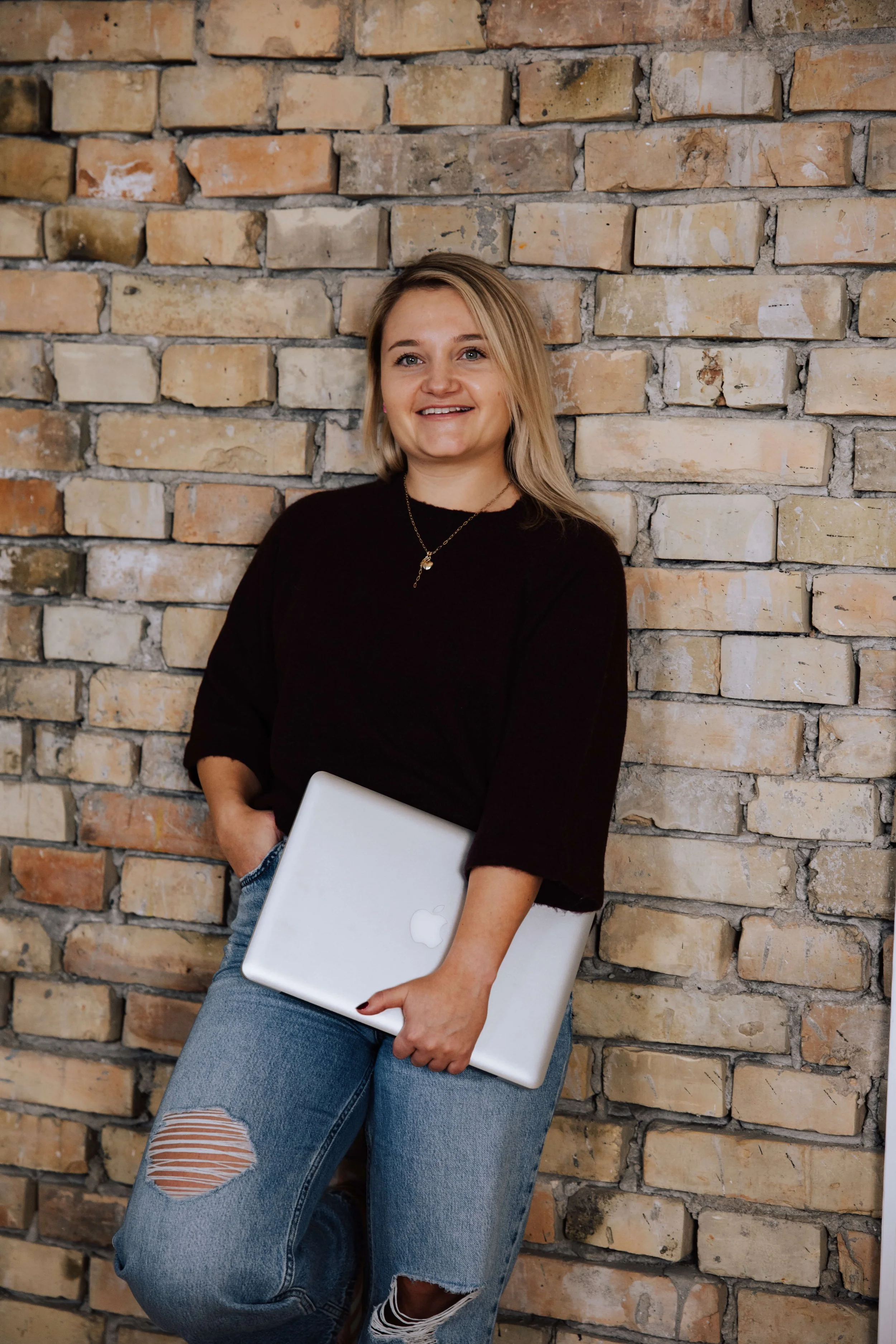 Eine lachende junge Frau mit blonden Haaren, schwarze Oberteil, ripped Jeans, hält einen silbernen MacBook in der Hand, steht vor einer Backsteinmauer.
