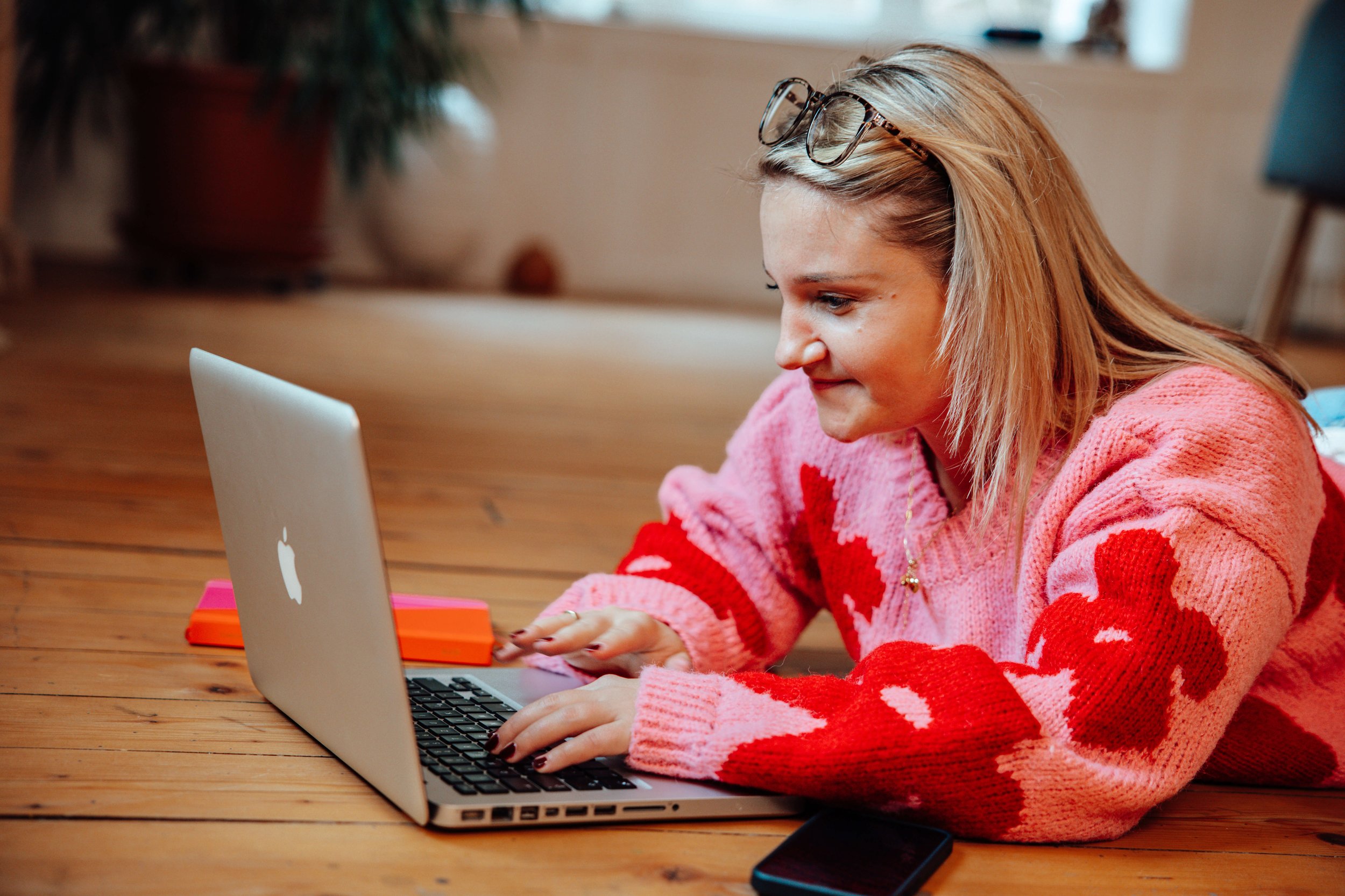 Junge Frau mit Brille auf Kopf arbeitet an einem Laptop auf Holztisch, trägt pinken Pullover mit roten Herzen, Handy neben Laptop, im Hintergrund Pflanzen und Möbel.