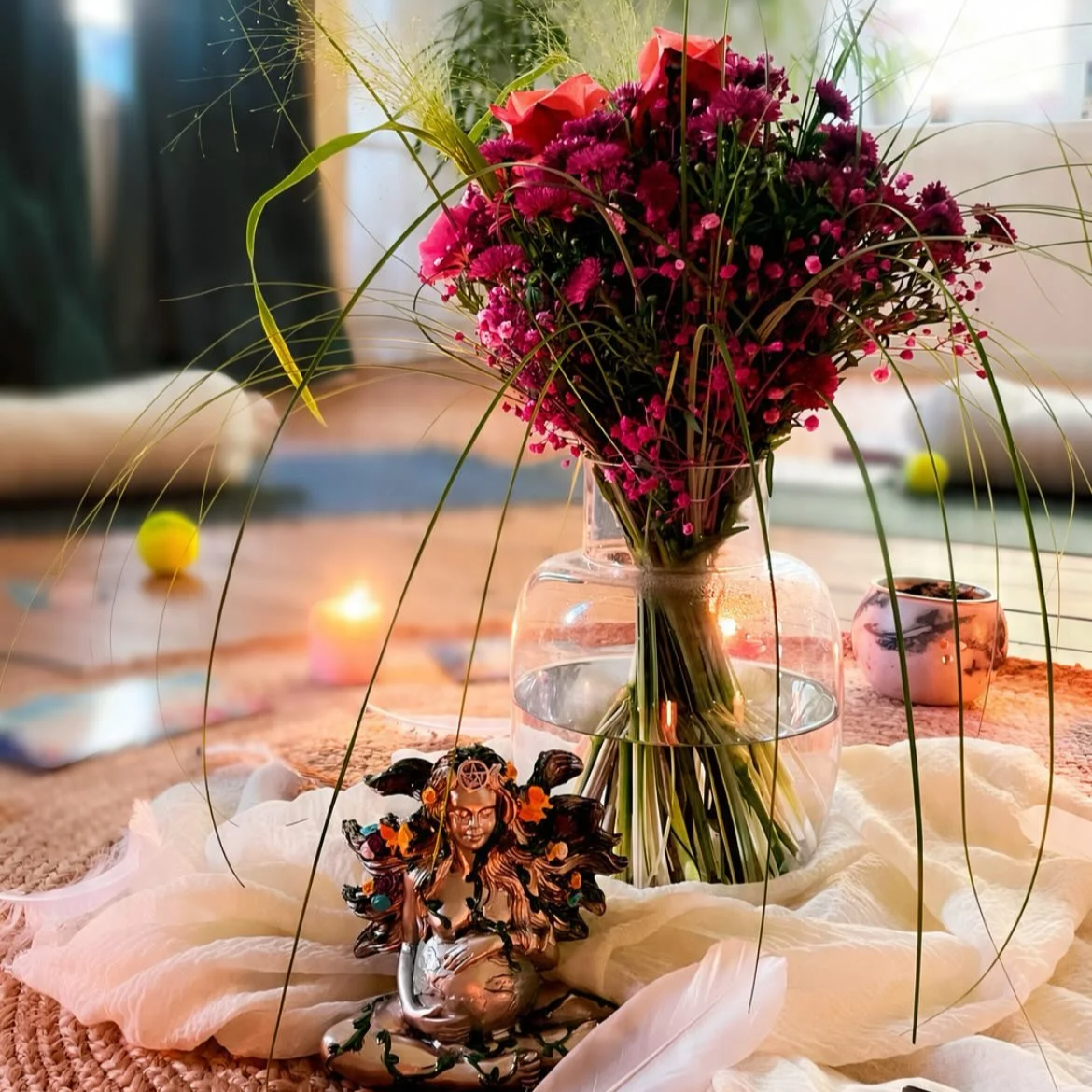 Blumenstrauß aus pinken und roten Blumen in einer Vasel mit Wasser, Kerze, kleine Figur einer Frau mit Flügeln und schwarzen Haaren, Keramikschale auf einem Tuch auf einem Tisch, im Hintergrund ein Tennisball und Kerzenlicht.