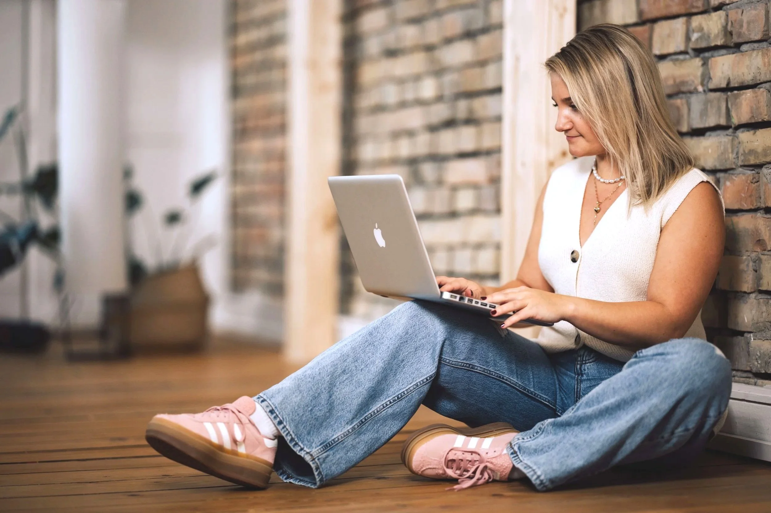 Junge Frau sitzt auf dem Boden vor einer Wand aus Backstein und benutzt einen MacBook-Laptop.