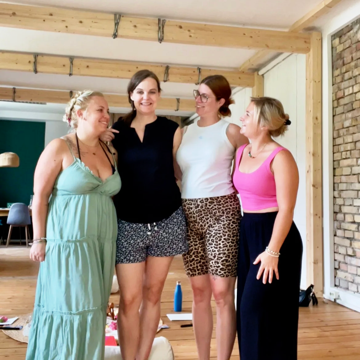 Vier Frauen stehen zusammen und lachen in einem Raum mit Holzdecke und Ziegelwand.
