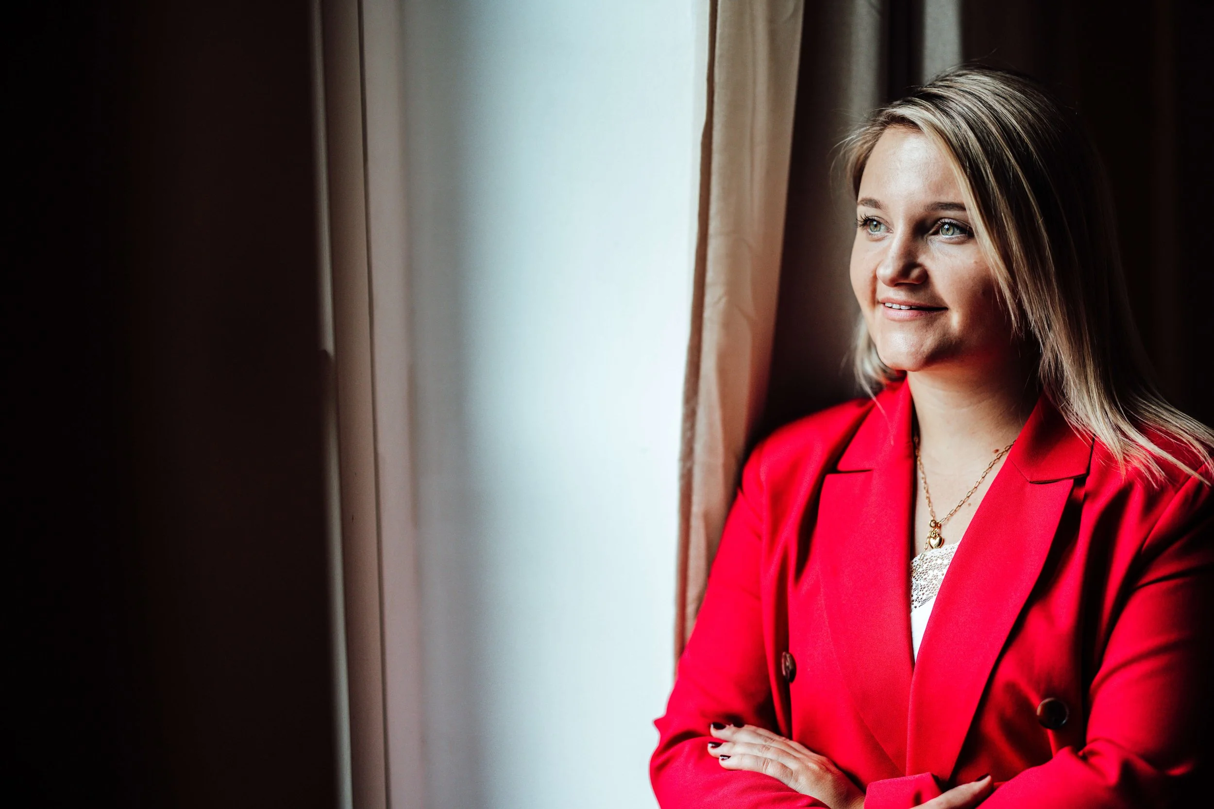 Eine lächelnde Frau mit blonden Haaren trägt eine rote Jacke und schaut aus einem Fenster.