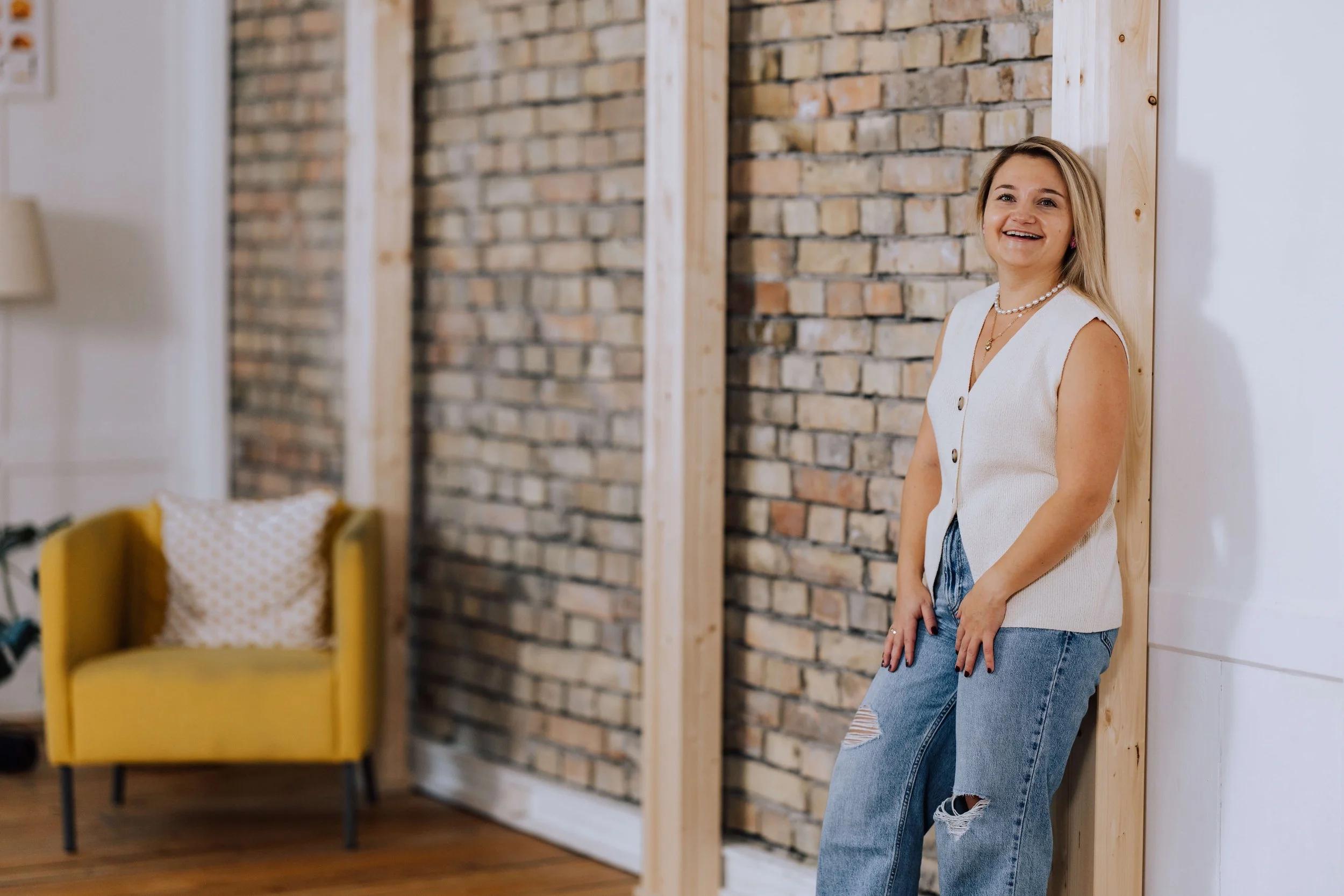 Junge Frau mit blonden Haaren lächelnd in einem rustikalen Raum, lehnt an Holzrahmen, im Hintergrund eine gelbe Sessel mit Polsterkissen.