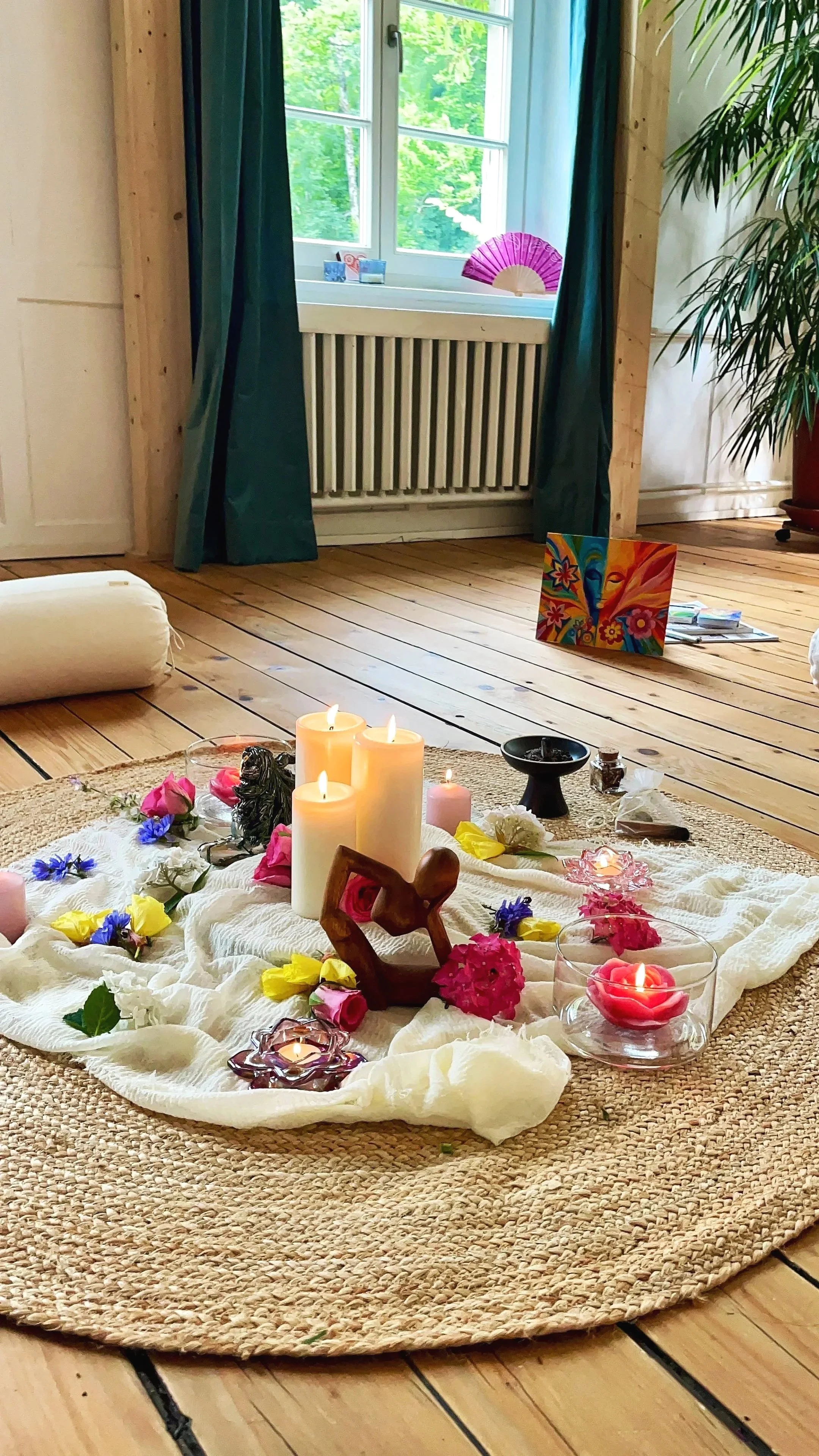 Ein mit Kerzen, Blumen und spirituellen Gegenständen gedeckter Altar auf einem runden Teppich in einem Raum mit Holzboden und großem Fenster.