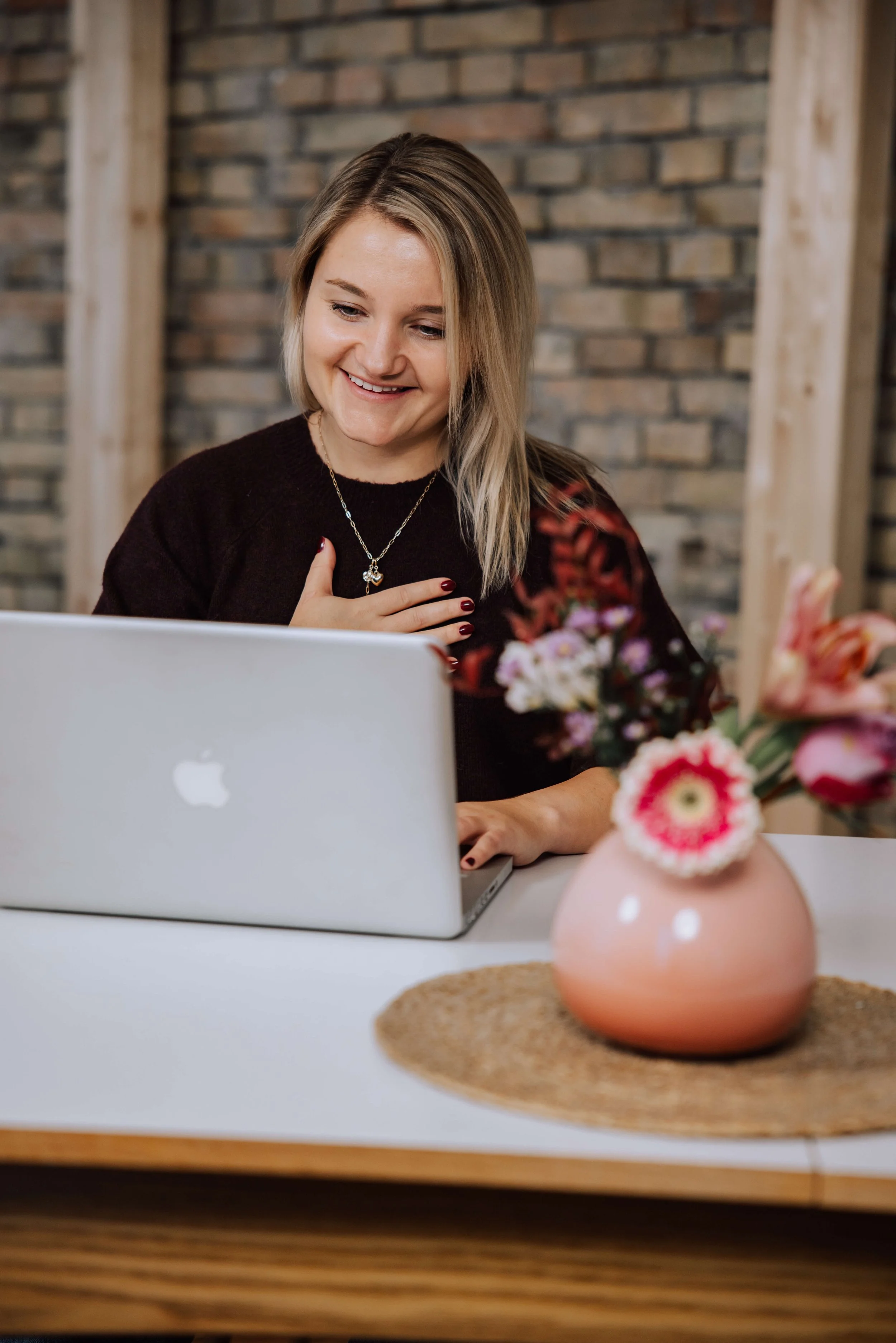 Eine lachende Frau sitzt an einem weißen Tisch vor einem Laptop, mit einer Hand an der Brust und schaut auf den Bildschirm, im Vordergrund eine Vase mit bunten Blumen.