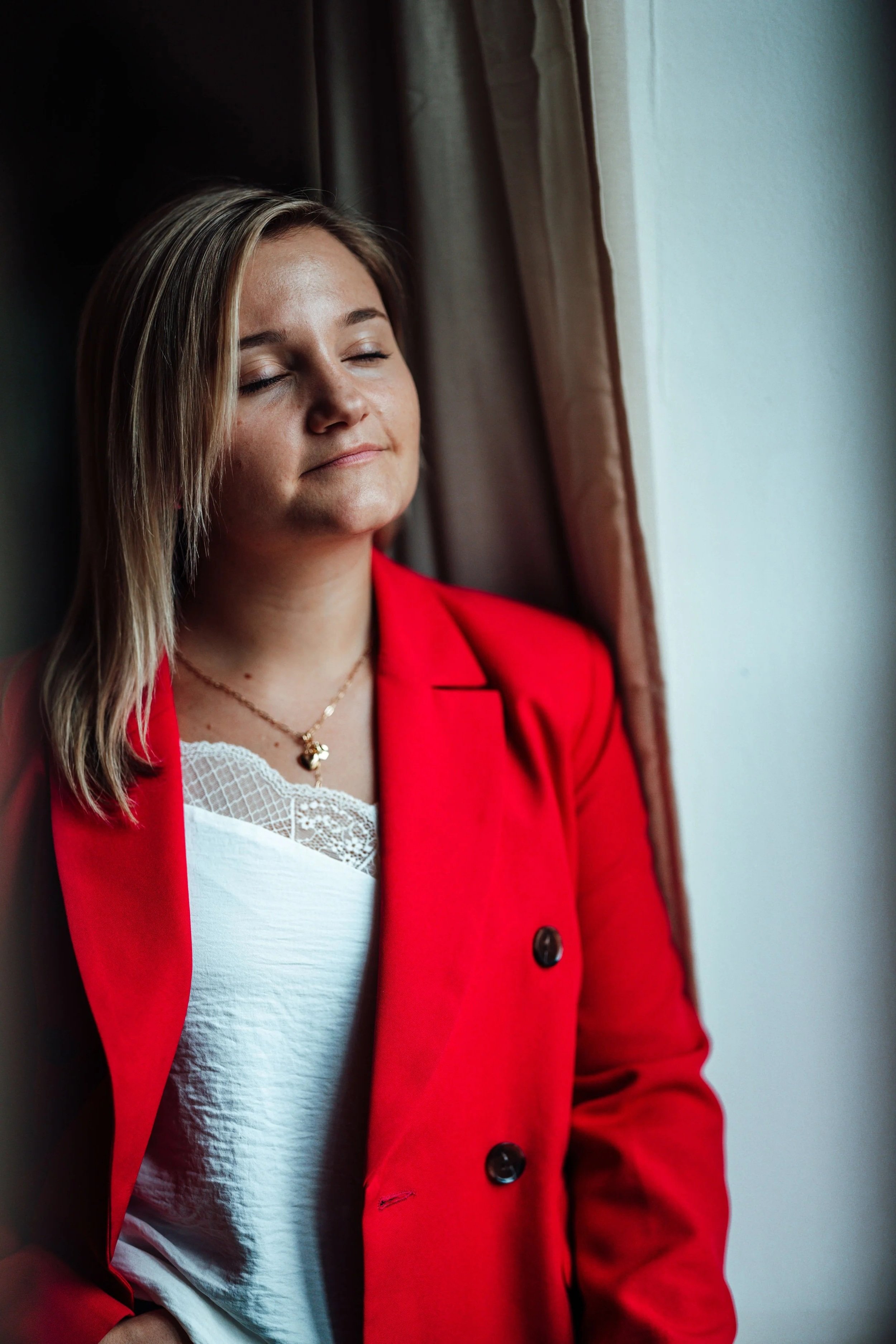 Eine junge Frau mit blonden, glatten Haaren, die ihre Augen geschlossen hat, trägt eine rote Jacke und ein weißes Top mit Spitzenmuster, steht vor einem Fenster mit einem braunen Vorhang.