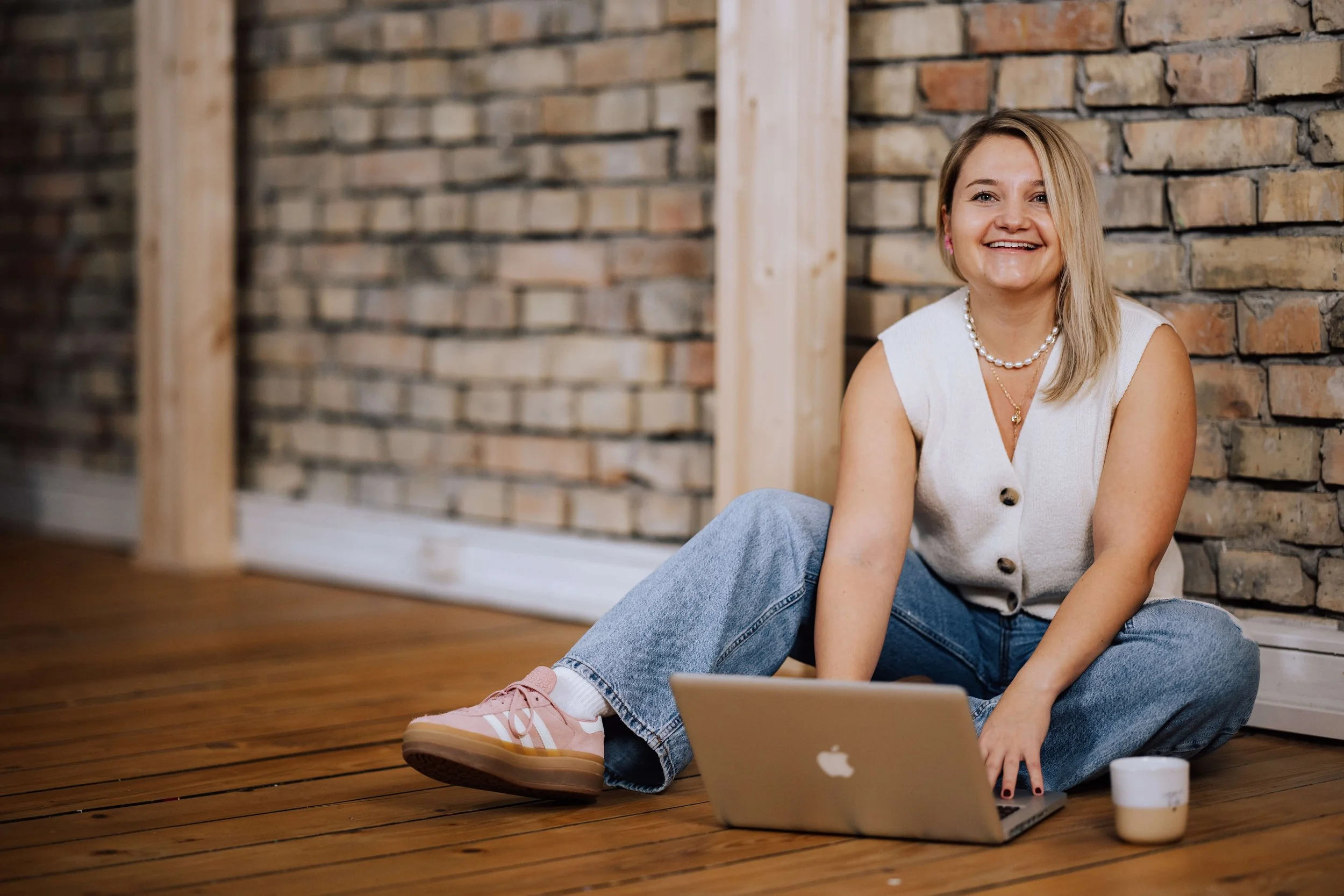 Eine junge Frau sitzt auf einem Holzfußboden vor einer Ziegelwand, arbeitet an einem Laptop, trägt eine weiße Weste, blaue Jeans, rosa Turnschuhe und hat eine Perlenkette. Es ist eine ungeordnete, entspannte Atmosphäre.