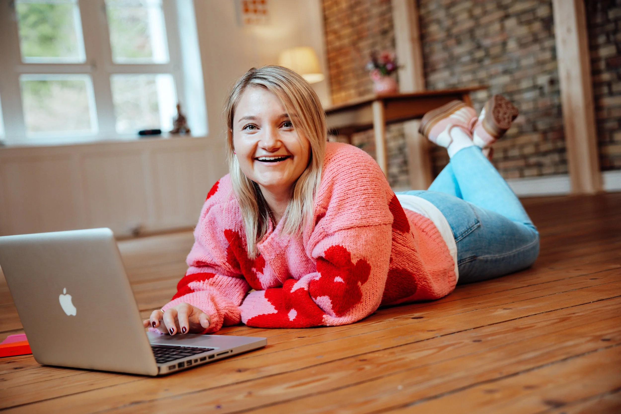 Lächelnde junge Frau liegt auf einem Holzboden vor einem Laptop, trägt einen pinken Pullover mit roten Herzen, in einem gemütlichen Raum mit Fenstern, einer Lampe und einer Blumen vase im Hintergrund.