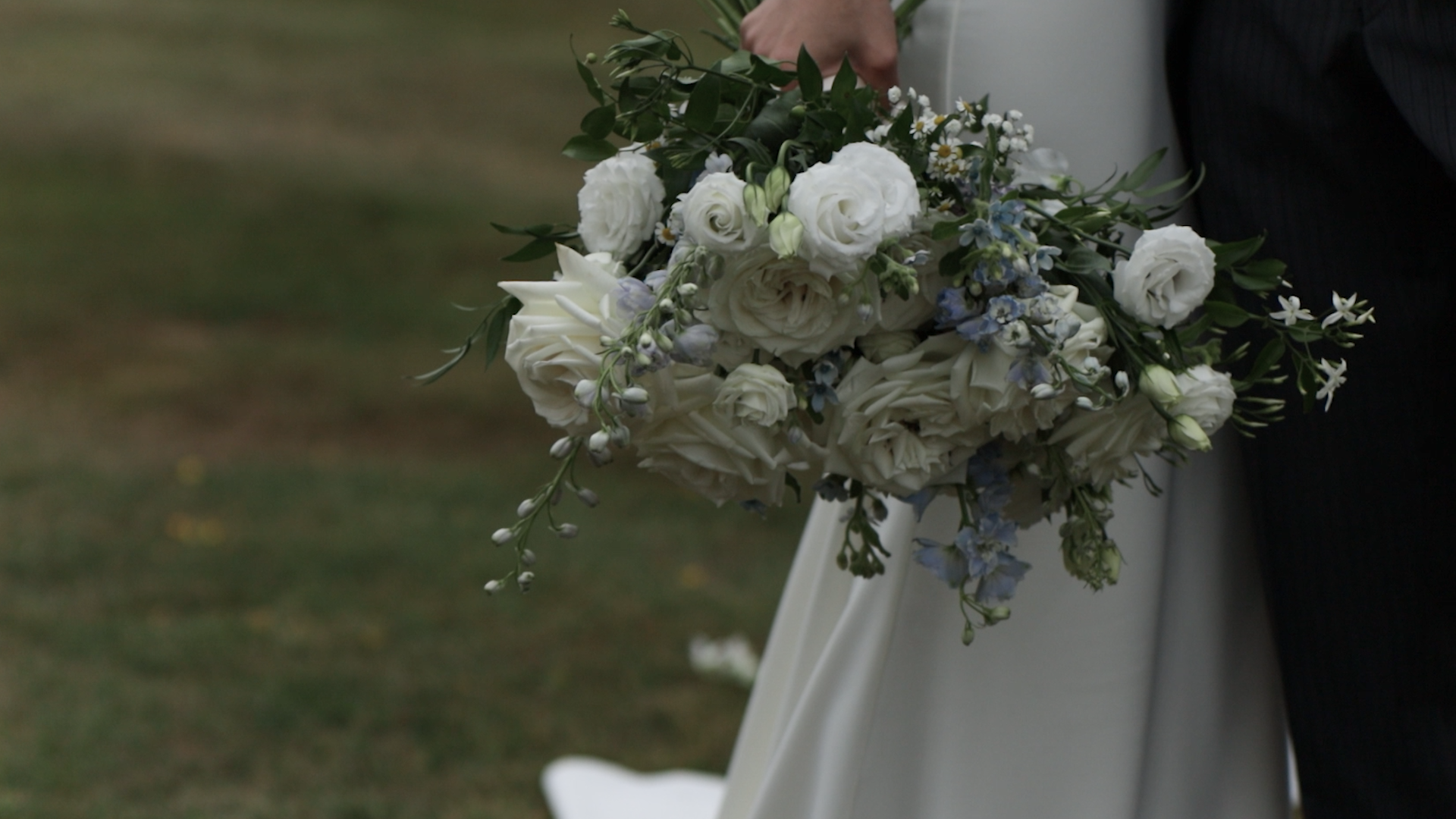 Captured by Natasha Gray Video, a bridal bouquet consisting of white roses, white lilies, small white daisies, and light blue flowers, held by a person in a chic wedding dress.