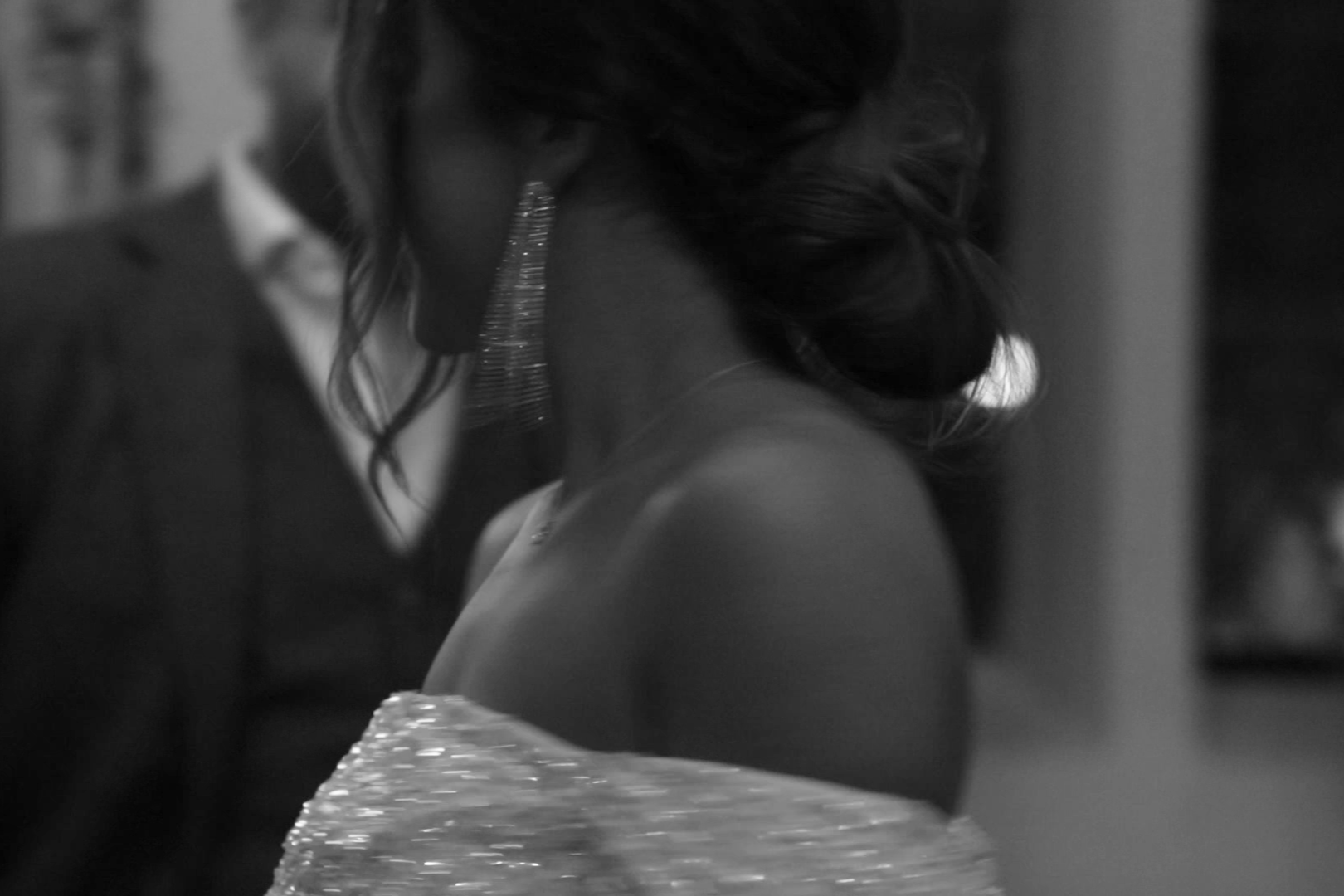 Black and white shot by London wedding videographer Natasha Gray Video. Close-up photo of a bride with curly hair wearing diamond earrings, a sparkly off-the-shoulder dress, and a necklace, turned slightly away from the camera.