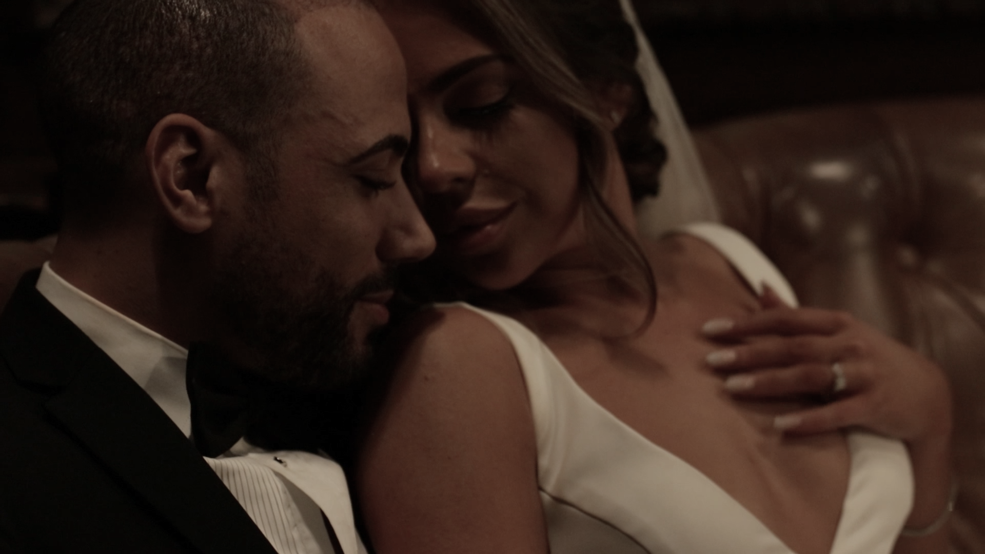 A couple in formal wedding attire sharing an intimate moment, sitting closely with their foreheads touching and eyes closed.