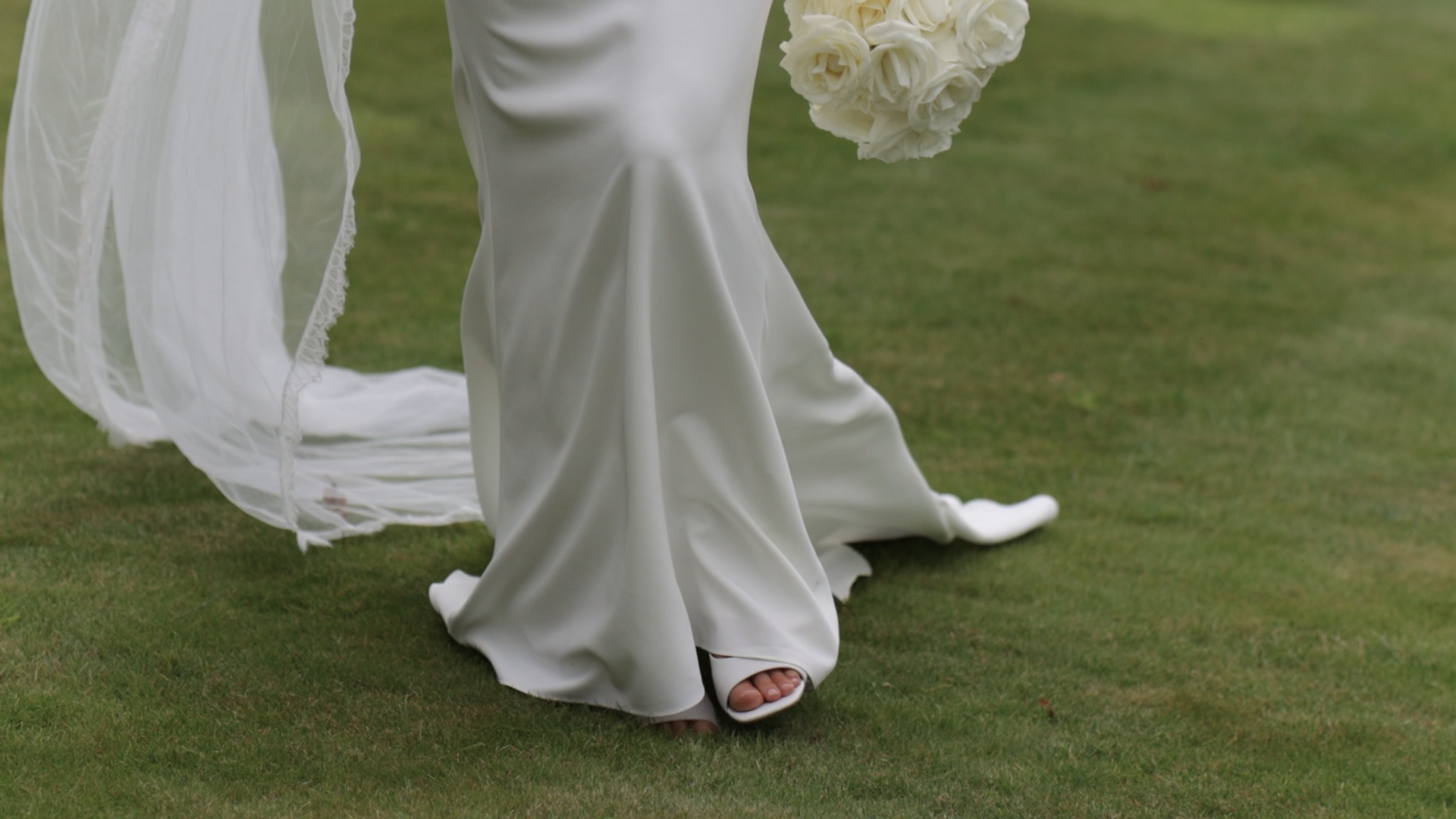 Close-up of a bride's white wedding dress and veil on grass, showing her feet in open-toed shoes, holding a bouquet of white roses.