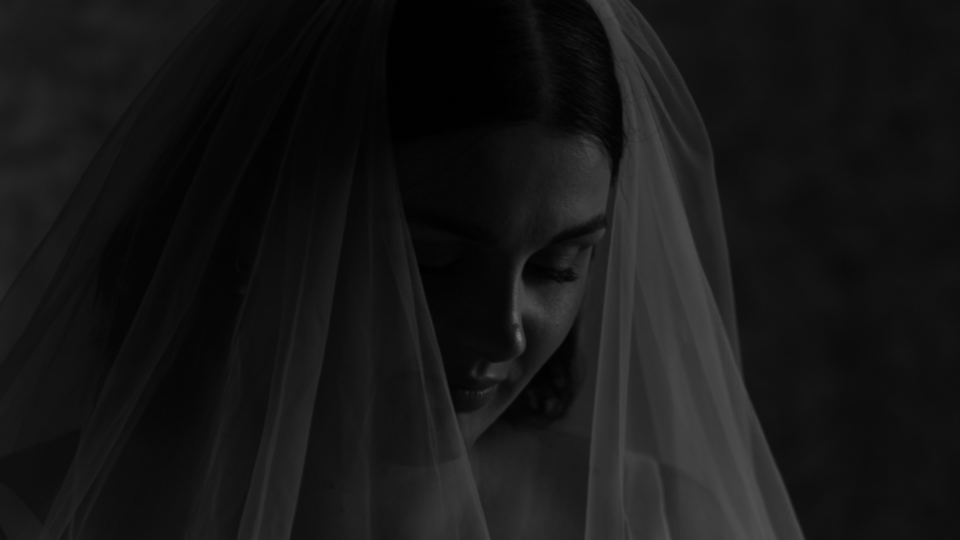 A close-up black and white photo of a woman with a veil over her head, looking down with a gentle expression.