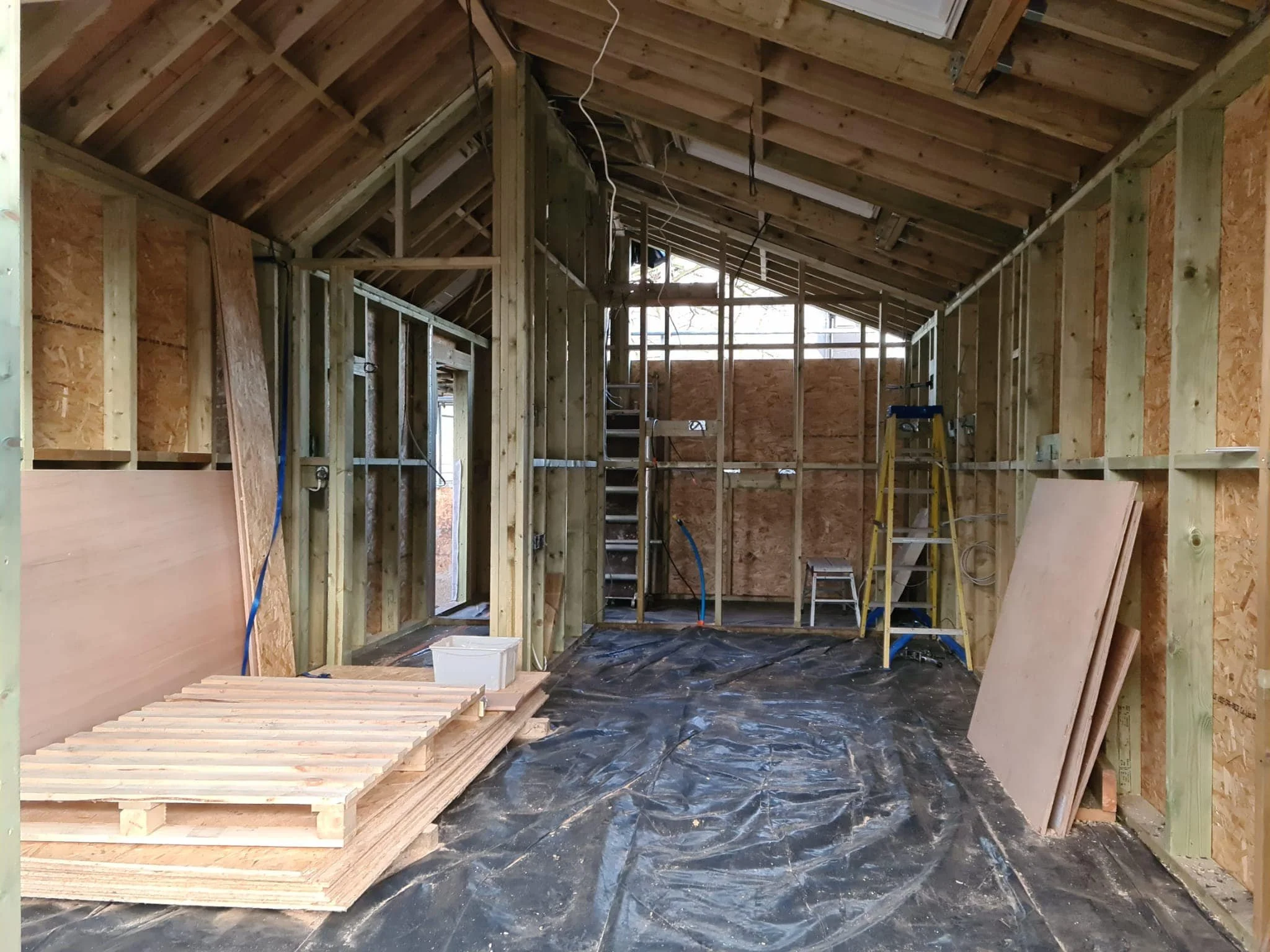 Interior of a house under construction with exposed wooden framing, a ladder, and construction materials on the floor.