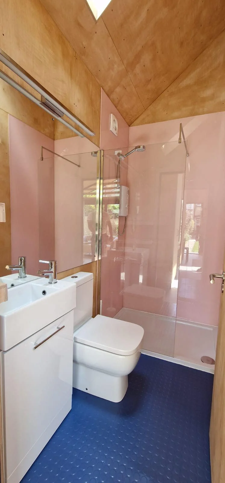 A small bathroom with a blue textured floor, a white toilet, a small white sink with modern faucets, and a glass-enclosed shower area with pink walls. Wooden ceiling and wall accents, a skylight, and a reflection of a person in the glass.