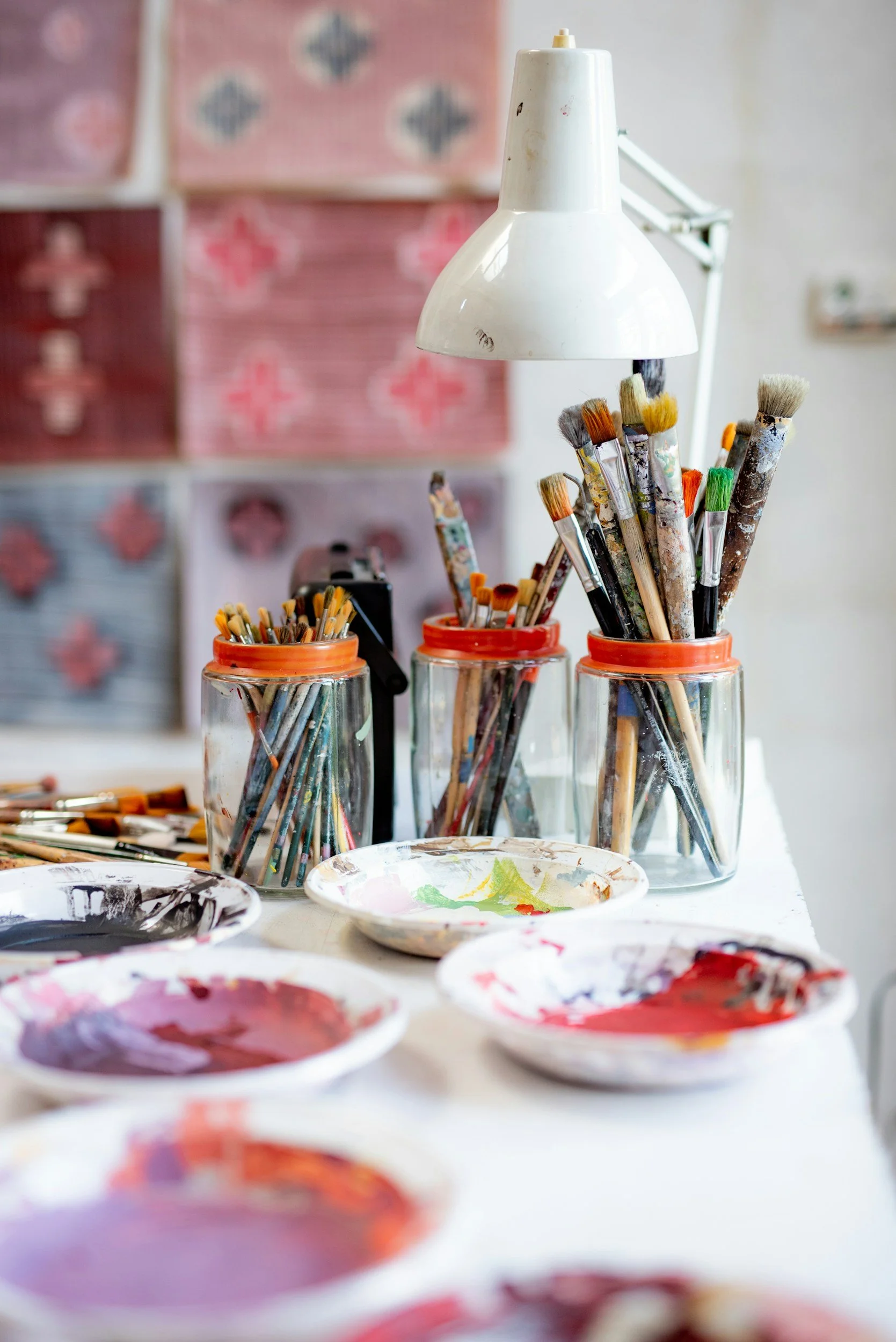 Artist's work area with jars of paintbrushes, paint palettes with mixed paints, and a white adjustable lamp overhead.