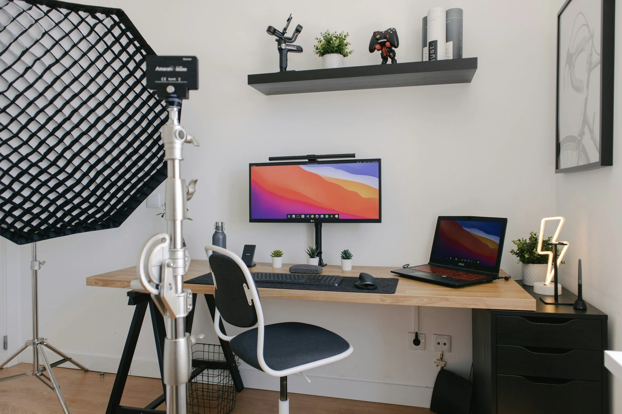 Home office setup with a wooden desk, desktop monitor, laptop, office chair, studio lighting, wall shelf with decor, and framed artwork.