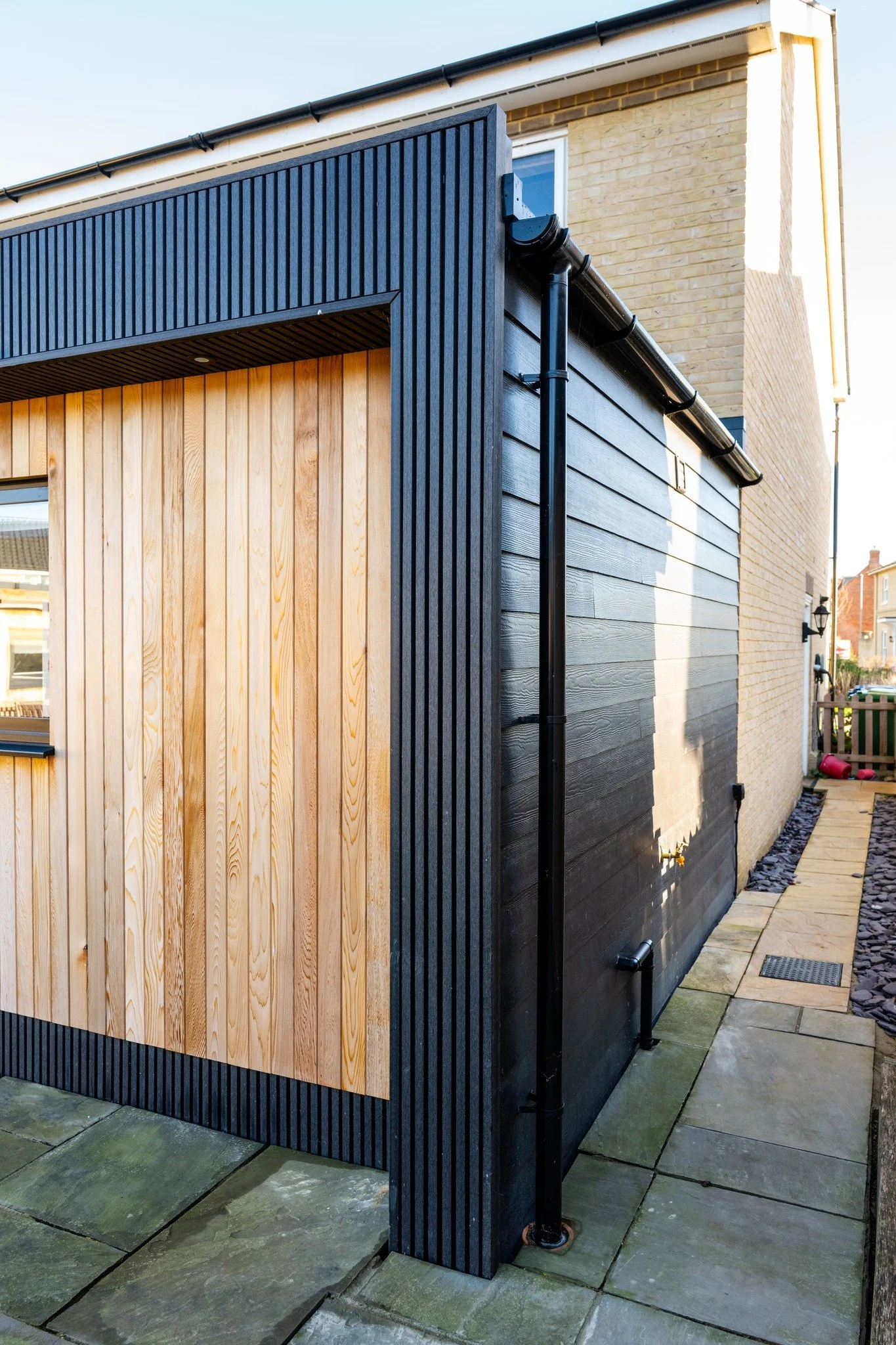 Exterior view of a modern building with black cladding and natural wood paneling, sewer pipes on the right, and a paved walkway.