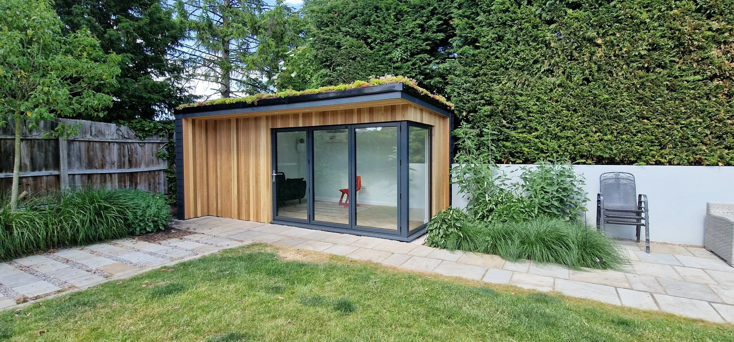 A modern garden room with wooden exterior and large glass sliding doors, situated in a backyard with a grassy lawn, paving stones, and surrounding greenery including bushes, trees, and a tall hedge. There are also some outdoor chairs and a wicker table on the patio.