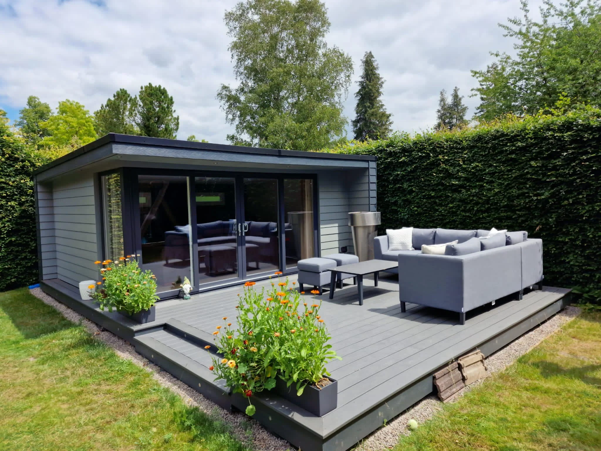 A modern backyard patio with outdoor seating, including a sectional sofa and a table. There are flower planters with orange and yellow flowers, a small wooden platform, and a small building with sliding glass doors, surrounded by greenery and trees.