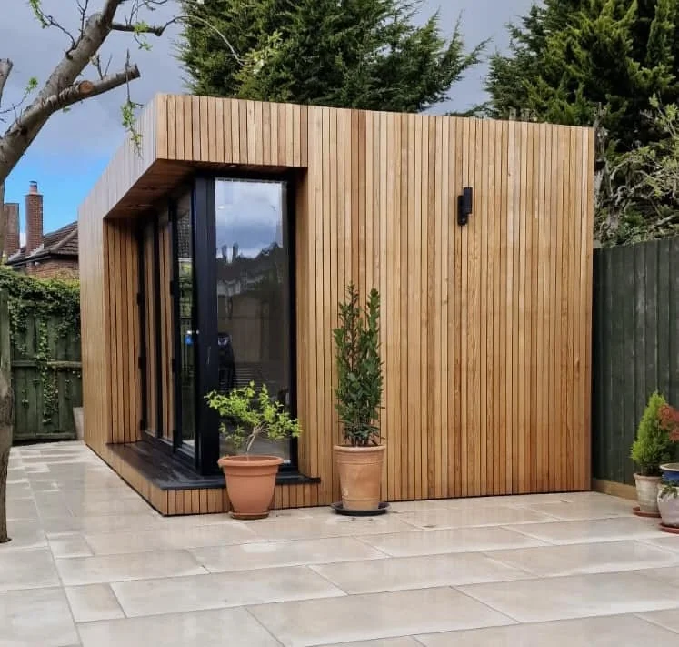 Small modern garden room with wooden siding, large glass sliding doors, and potted plants outside, in a backyard with a paved patio and a green fence.