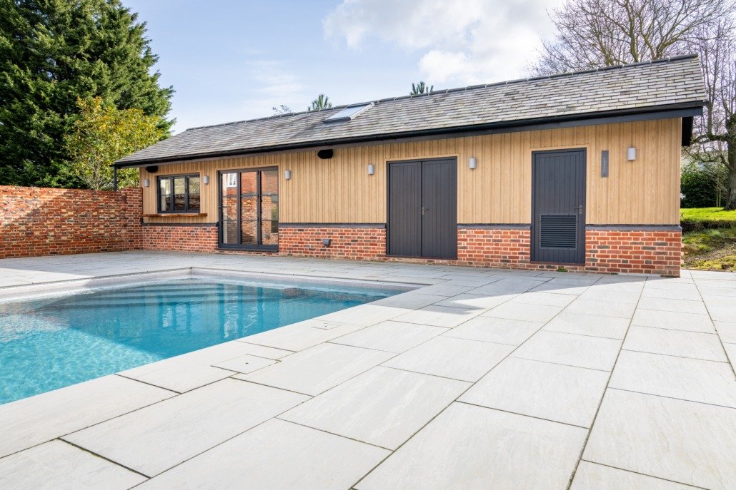 A backyard with a swimming pool, a wooden shed with black doors, and a surrounding concrete patio with a brick wall on one side.