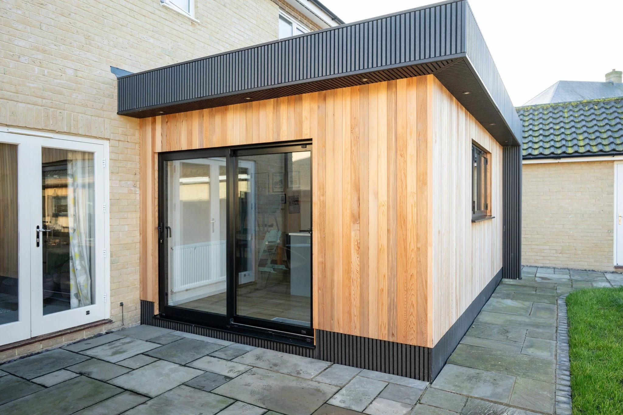 Exterior view of a modern backyard addition with sliding glass door, wooden siding, and a black metal overhang, next to a brick house.
