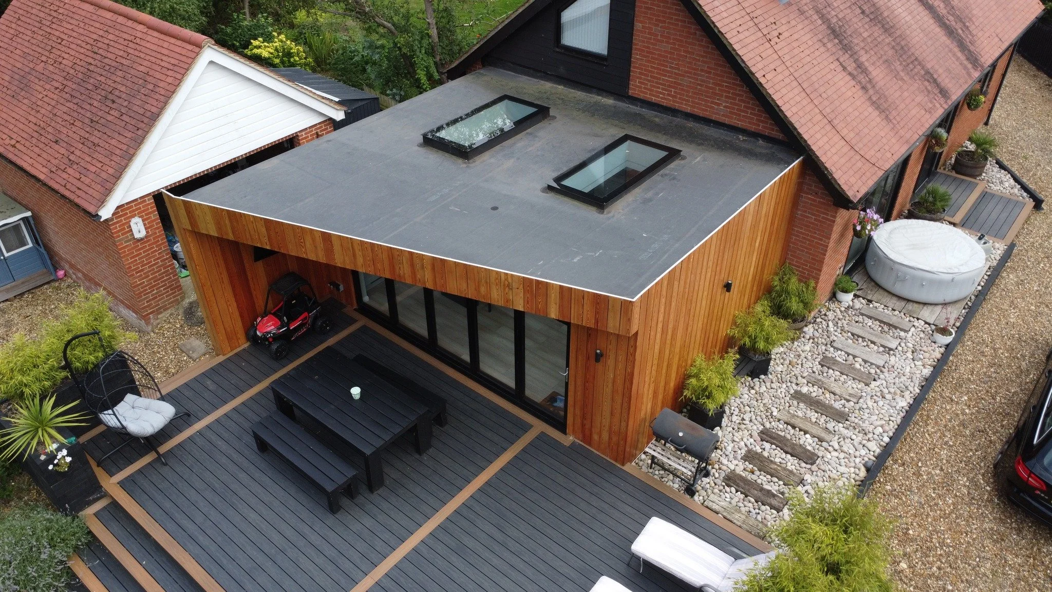Aerial view of modern backyard with wooden deck, black outdoor dining table with benches, small outdoor sofa, hot tub, potted plants, gravel pathway, and neighboring houses with red tile roofs.