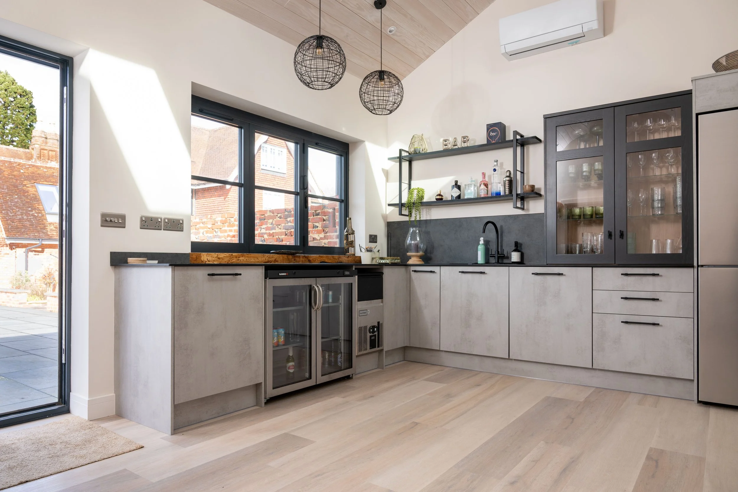 Modern kitchen with black-framed windows, light gray cabinets, black handles, exposed brick wall outside, and floating shelves with bottles and decor, featuring a wooden countertop and pendant lights.
