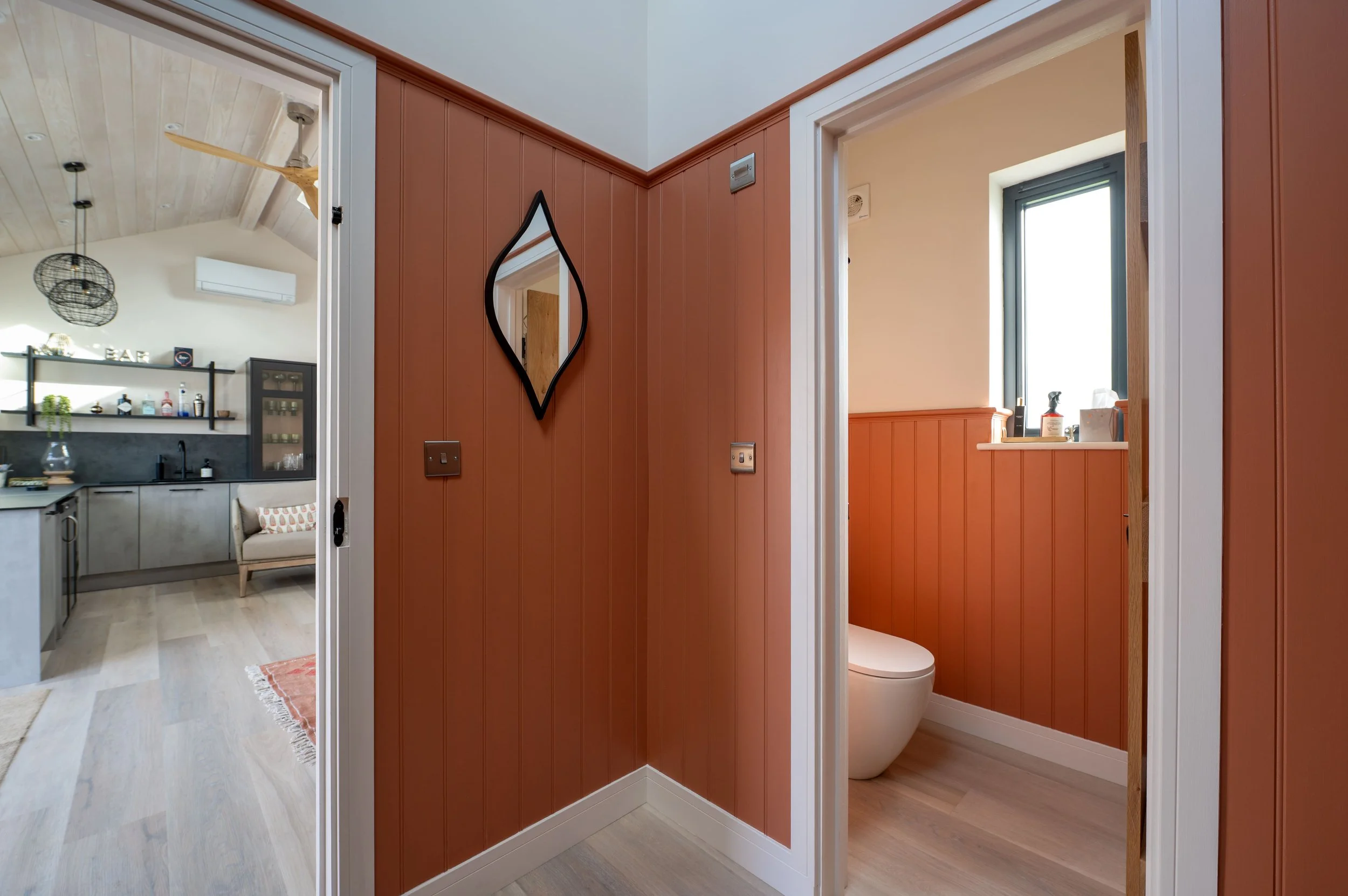 Interior view of a small bathroom with a modern toilet and a window, next to a reddish-brown wall with a black diamond-shaped mirror on it, in a home.