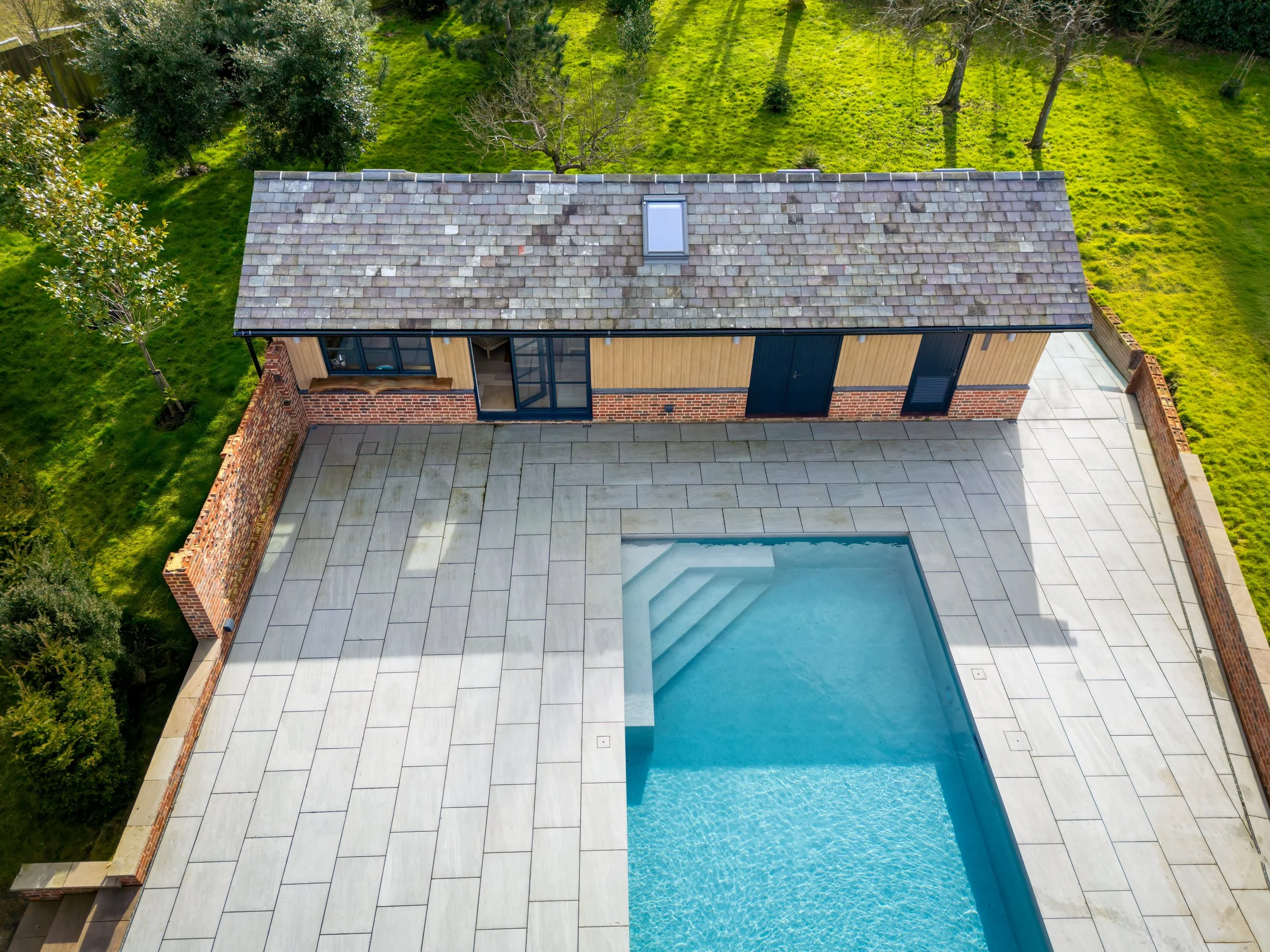 Aerial view of a house with a tiled roof, brick and wood exterior, and a backyard with a swimming pool surrounded by large paving stones, enclosed by a brick wall. The backyard has green grass and trees.