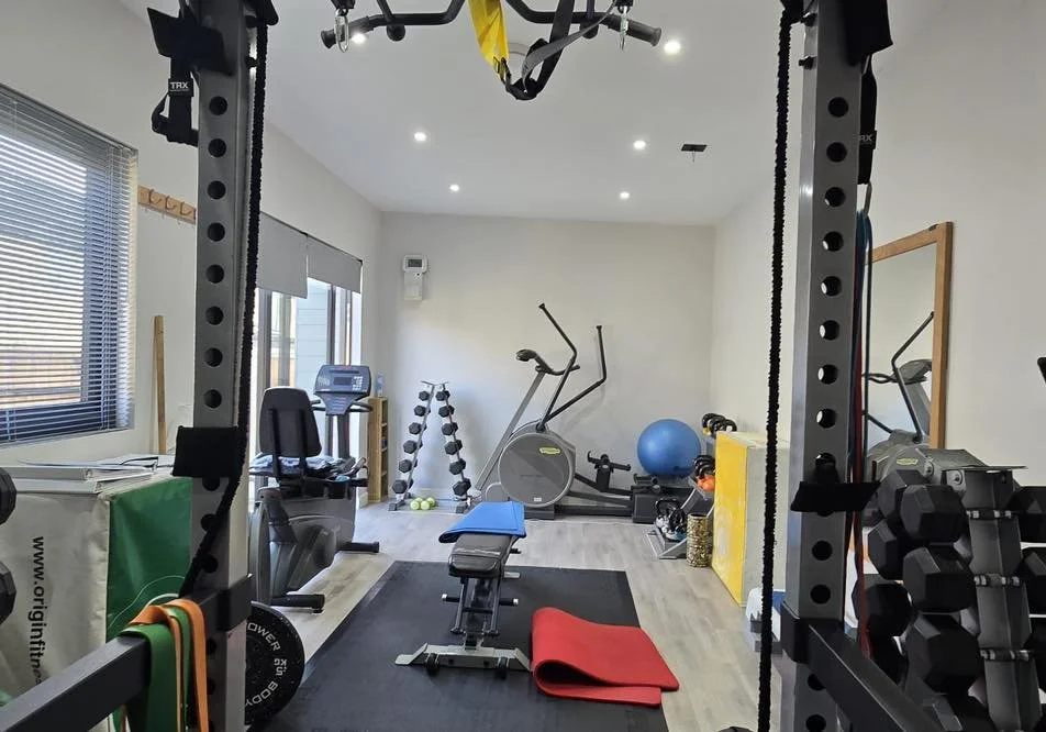 Fitness gym with exercise equipment including a treadmill, stationary bike, dumbbells, stability ball, and weight racks, with a mirror on the wall and windows letting in natural light.