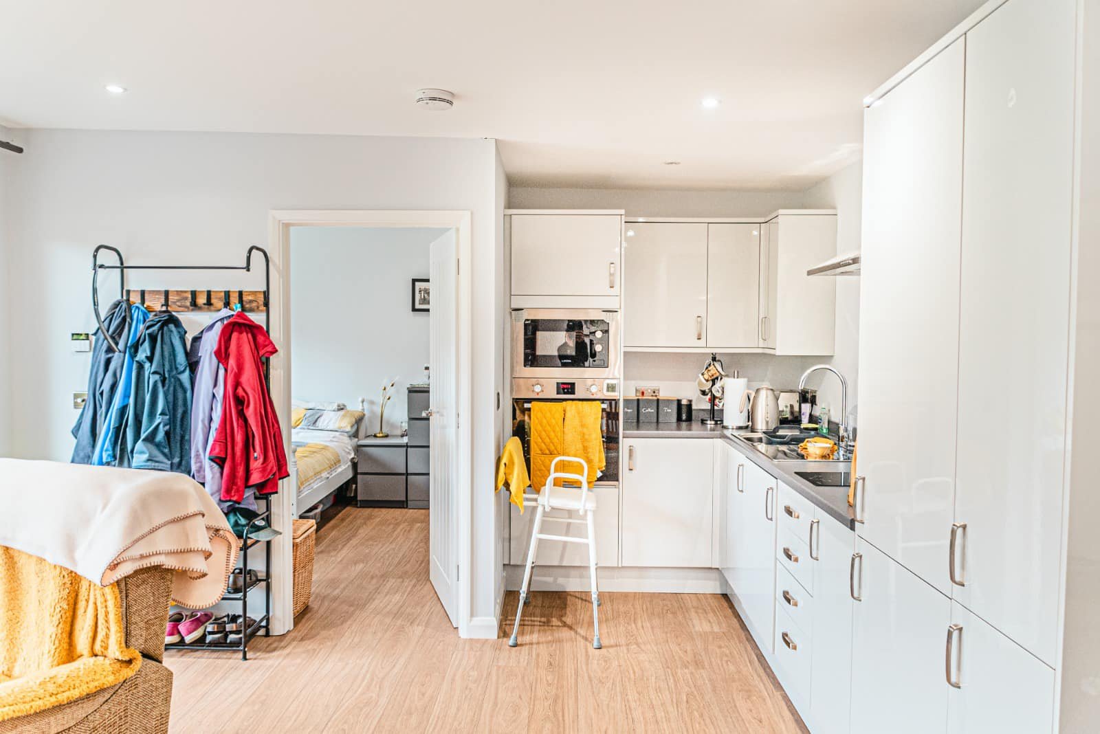 Kitchen and bedroom area in an apartment. The kitchen has white cabinets and a black countertop with various appliances. In front of the kitchen is a white step ladder with a yellow towel hanging on it. To the left is a bedroom with a bed, nightstand, and a clothing rack with jackets and clothes.
