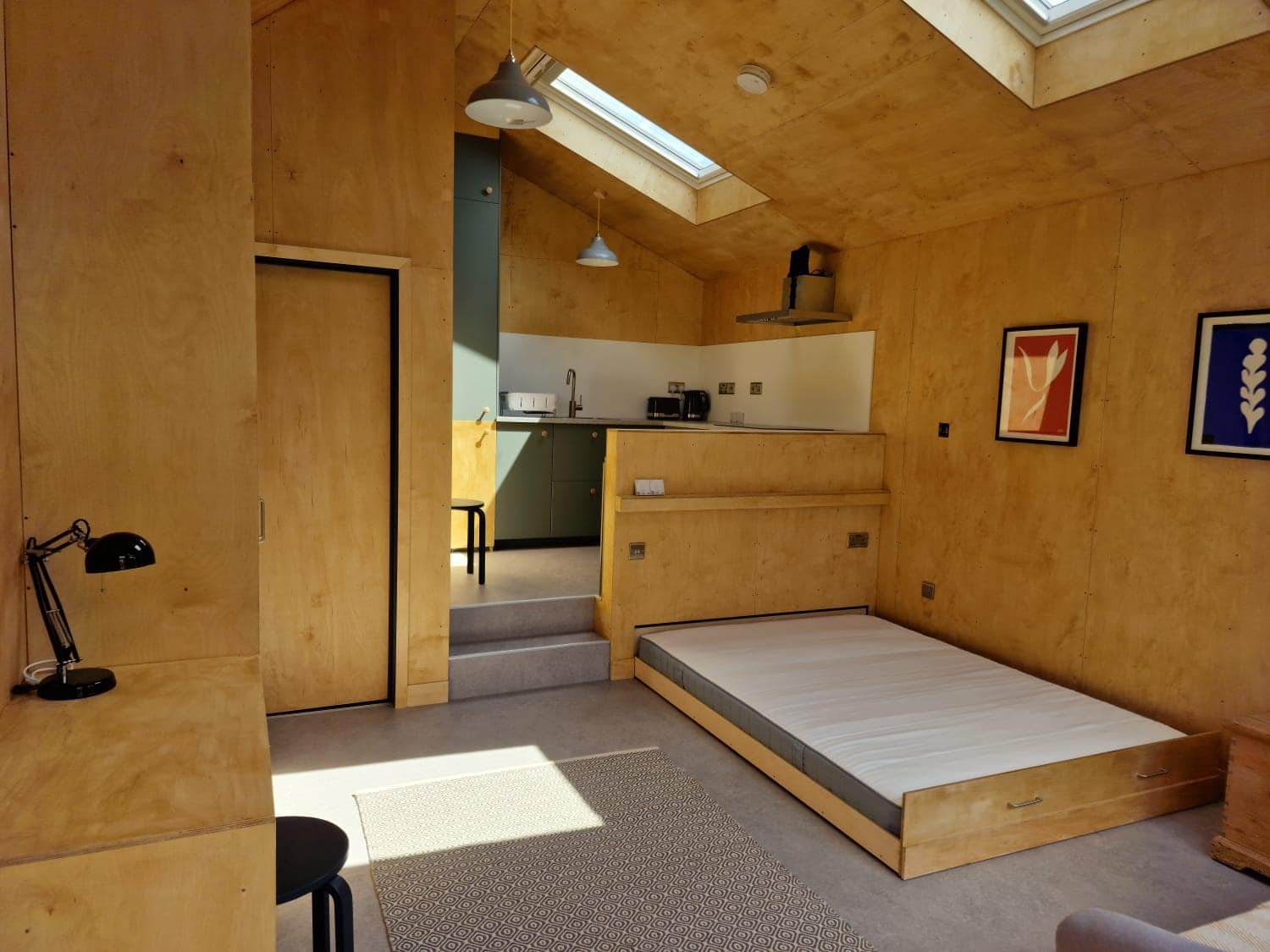 A minimalist room with wooden walls and ceiling, skylight windows, a lowered bed frame with a mattress, a desk with a black lamp, colorful framed art, and a small open kitchen area in the background.