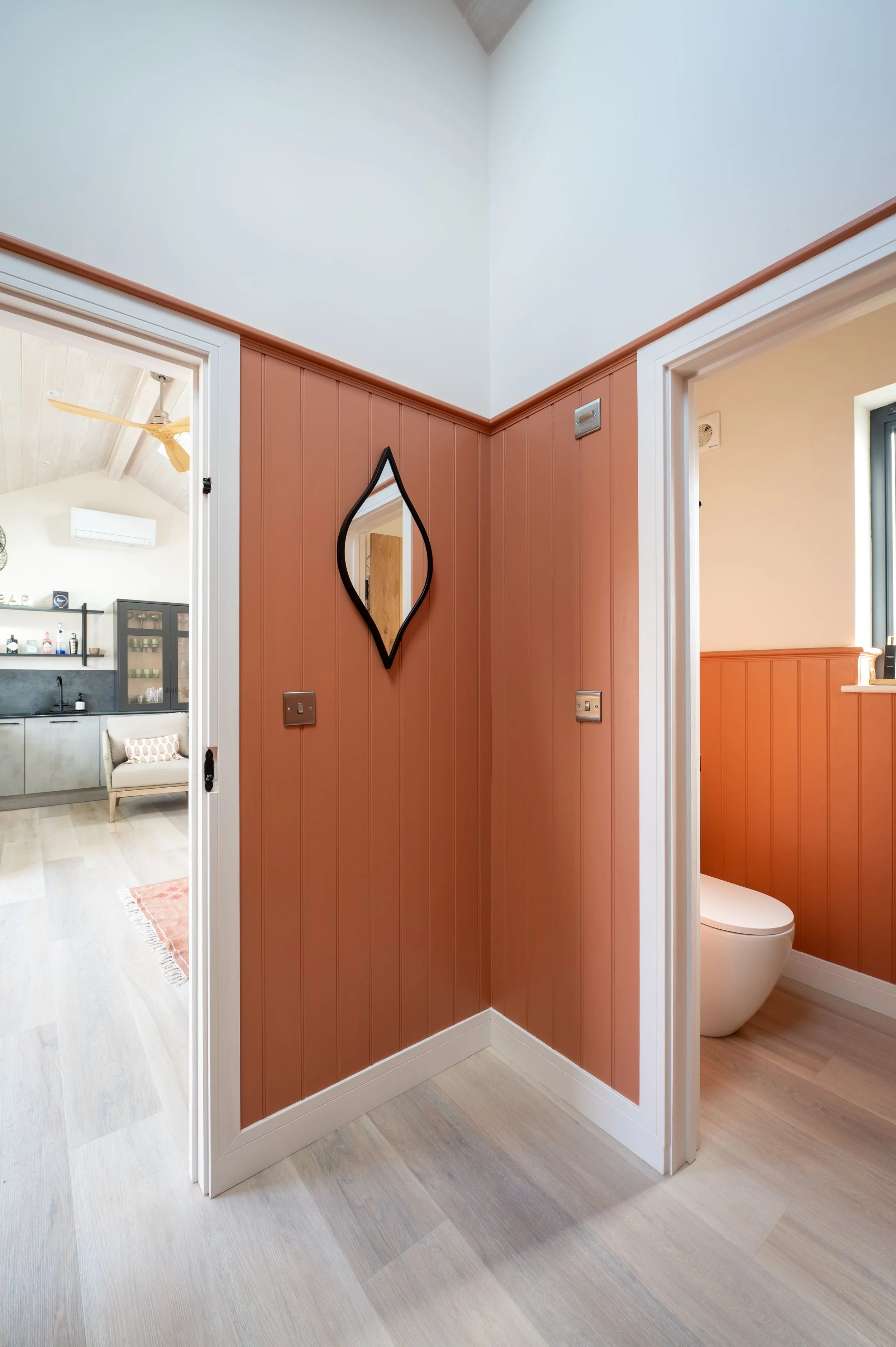 A hallway corner with two doorways; one leads to a bedroom or living area, the other to a bathroom with a toilet. The walls are wood-paneled painted in a brick red color, with a black, uniquely shaped mirror hanging on the wall.