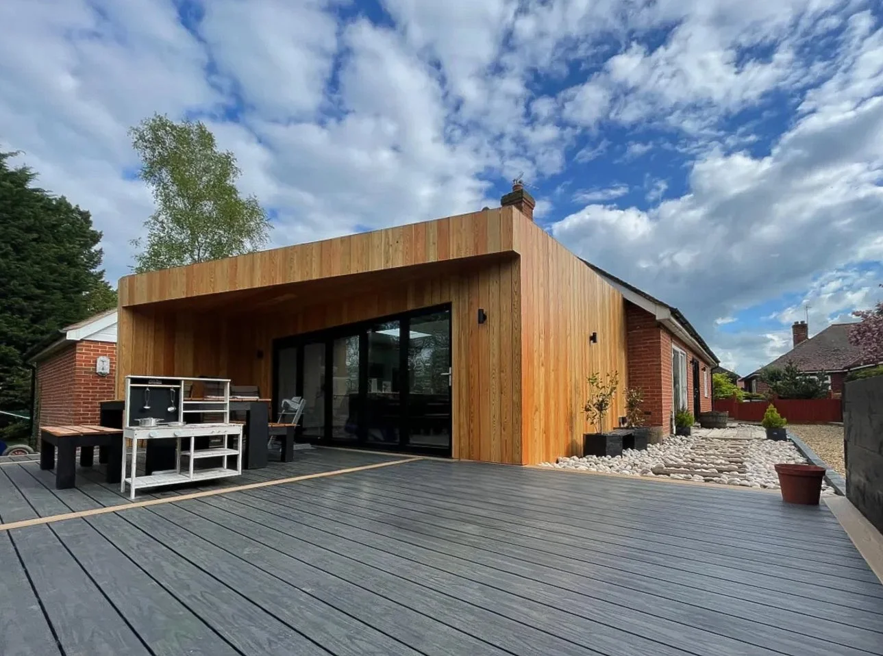 Modern house with a wooden exterior and large sliding glass doors, patio area with outdoor furniture, potted plants, and a cloudy sky.