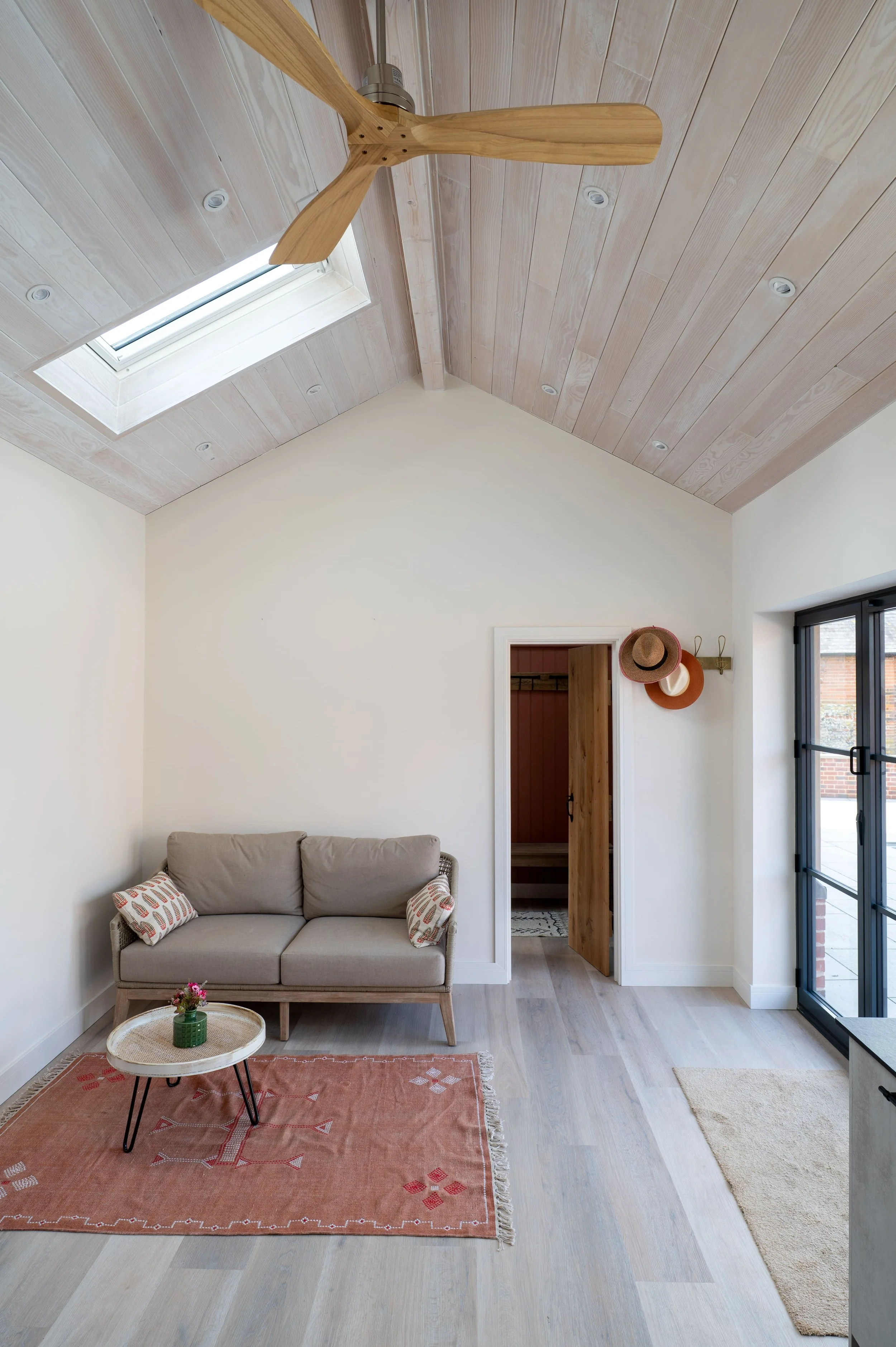 Living room with a beige couch, small round coffee table with a plant, pink rug, light wood flooring, sliding glass door, wooden ceiling with skylight, and hats hanging on a wall hook.