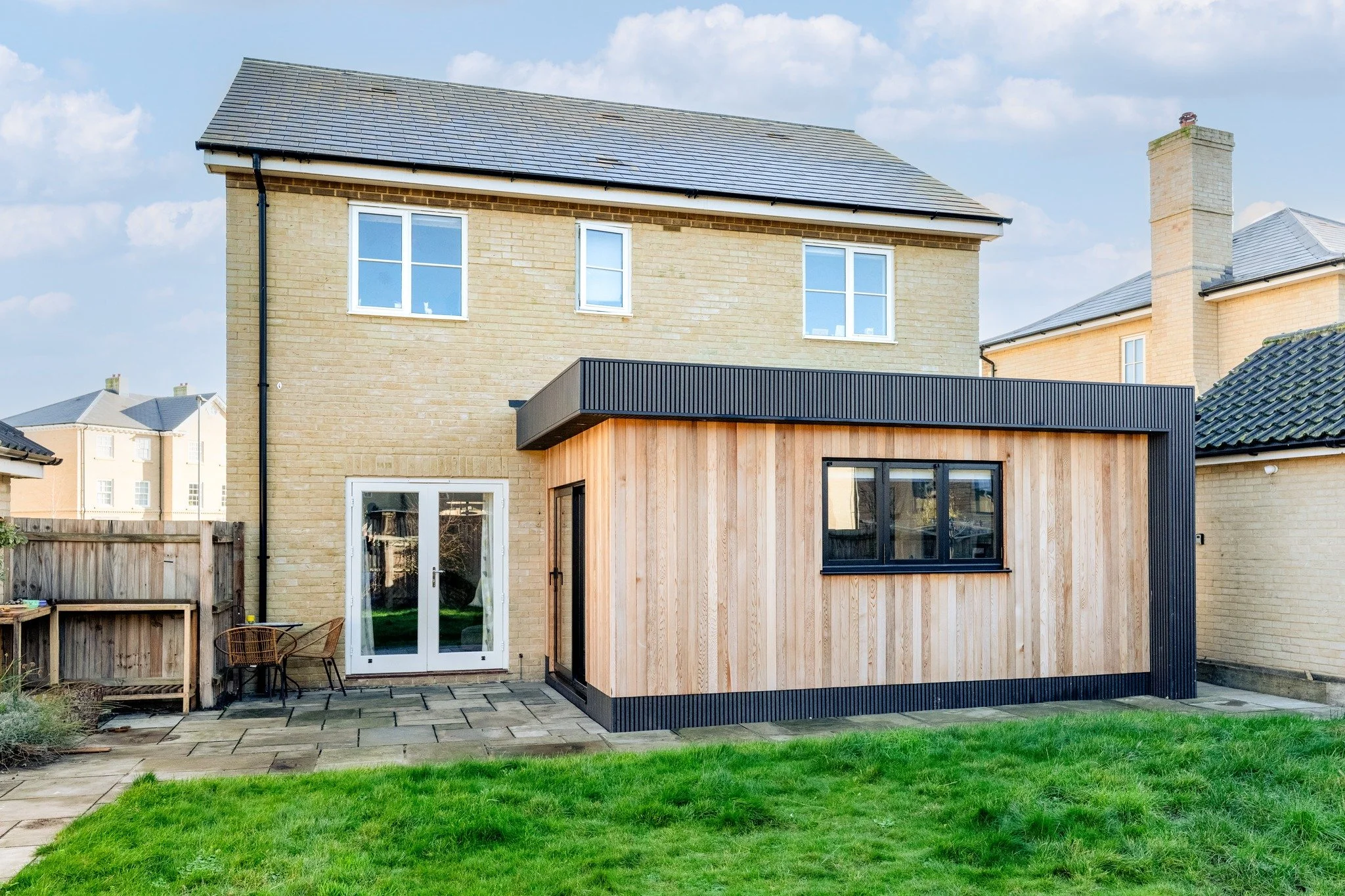 Backyard view of a two-story house with a modern detached wooden extension, green lawn, patio area, and neighboring houses in the background under a partly cloudy sky.