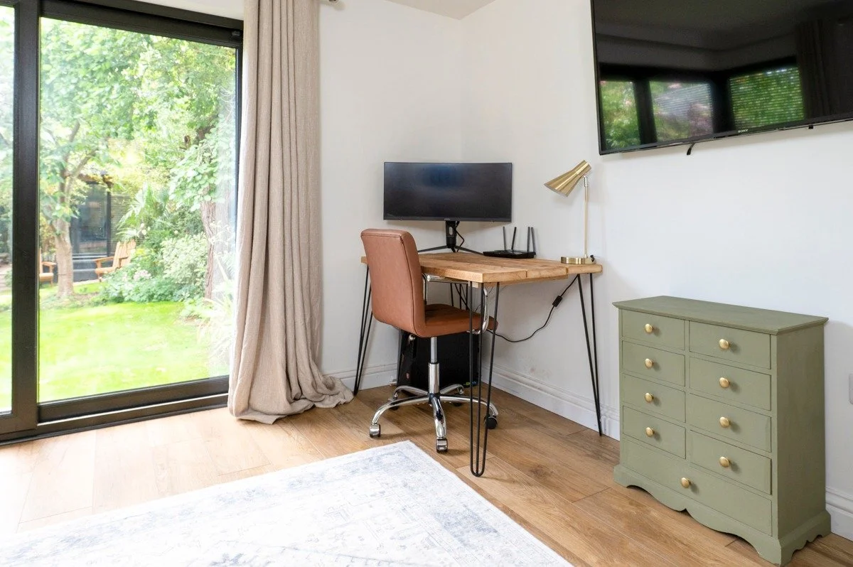 Home office with a computer desk, a brown chair, a desk lamp, a cabinet, and a large TV, with sliding glass doors leading to a garden.