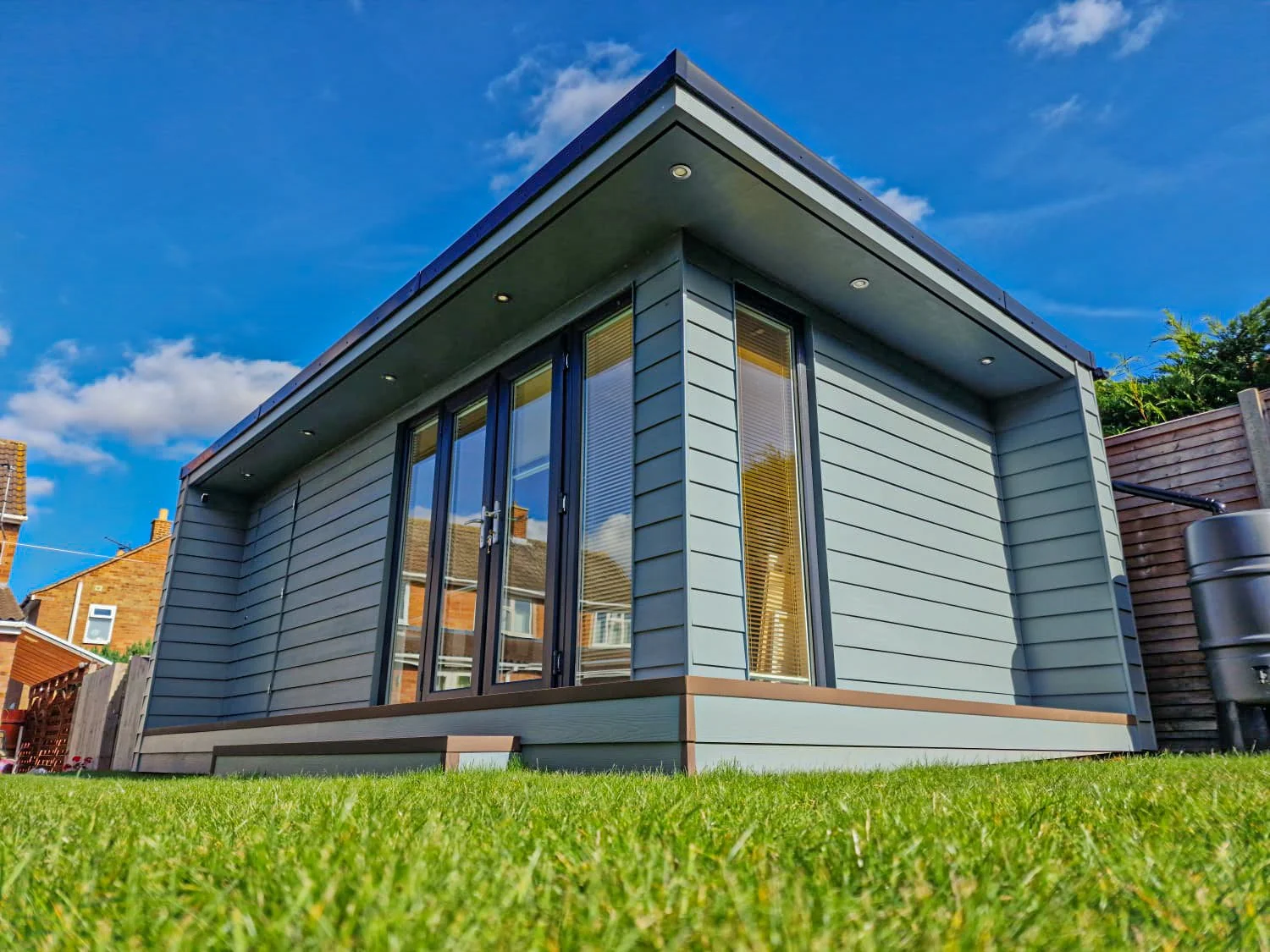 Modern, grey, single-story house with large glass doors and windows, surrounded by a green lawn and a wooden fence, under a partly cloudy blue sky.