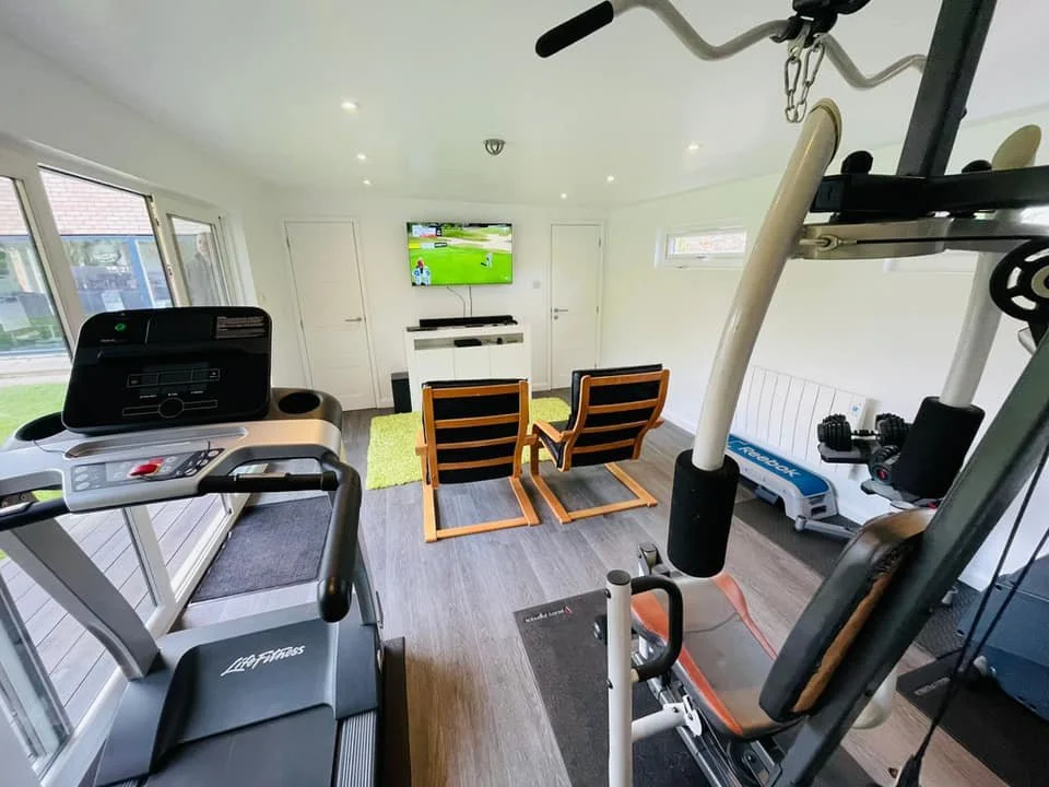 A home gym with a treadmill, weightlifting equipment, and a TV showing a golf game. There are two wooden chairs and a small green rug, with sliding glass doors on one side and a window on the other.