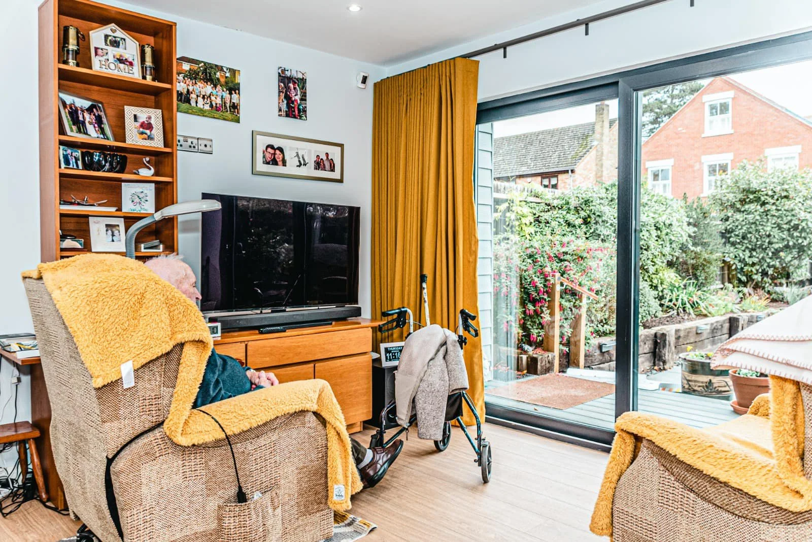 Living room with an elderly person sitting in a chair covered with a yellow blanket, near a walker, in front of a TV, with a sliding glass door opening to a garden outside.