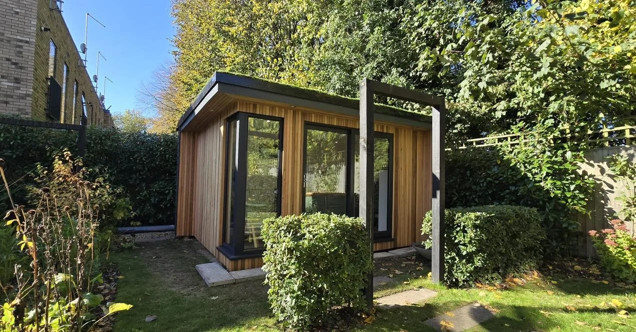 Small modern wooden backyard structure with glass doors, surrounded by greenery and bushes, with a blue sky overhead.