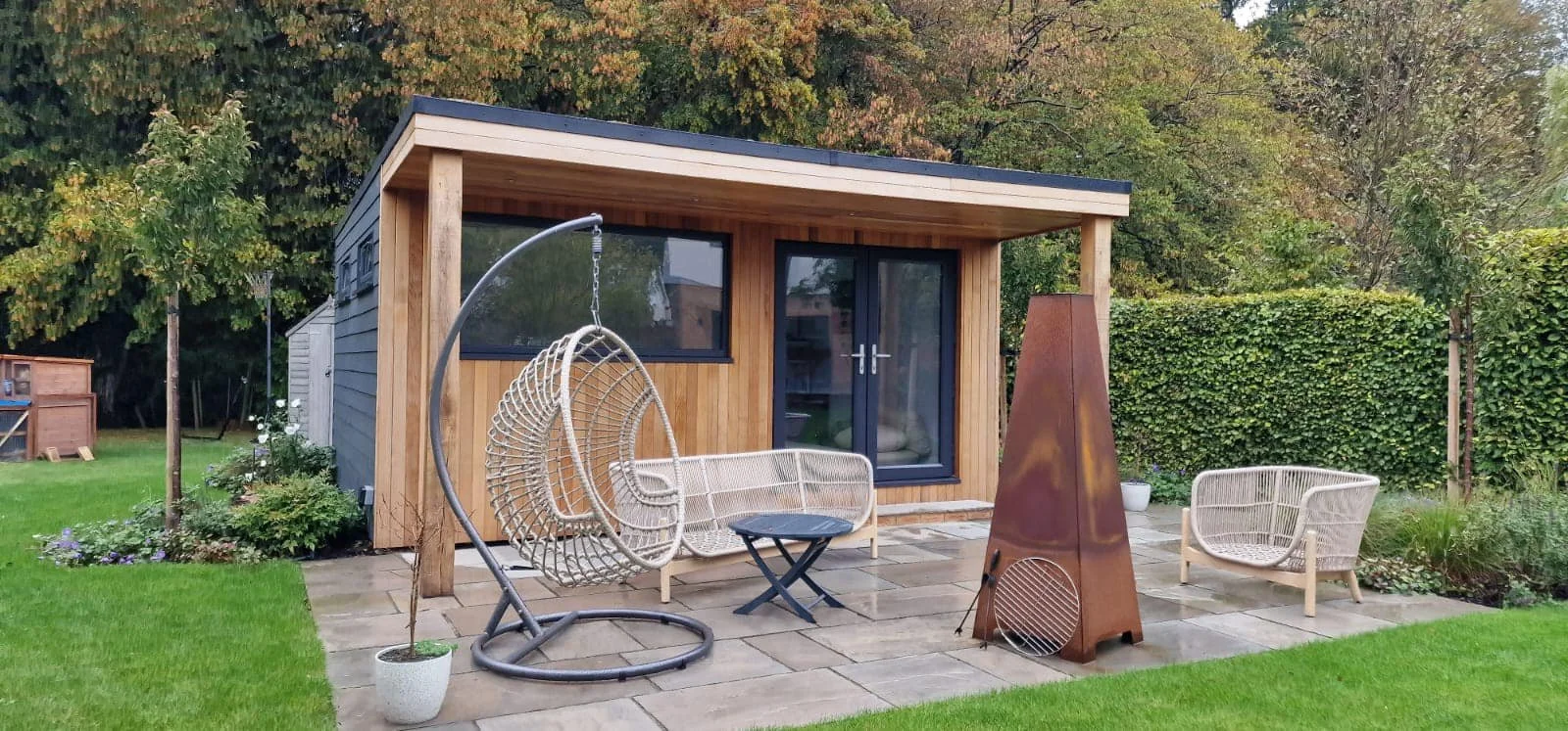 A backyard patio with two wicker chairs, a black metal table, a hanging egg chair, a modern outdoor heater, lush green grass, trees, bushes, and a small wooden shed in the background.