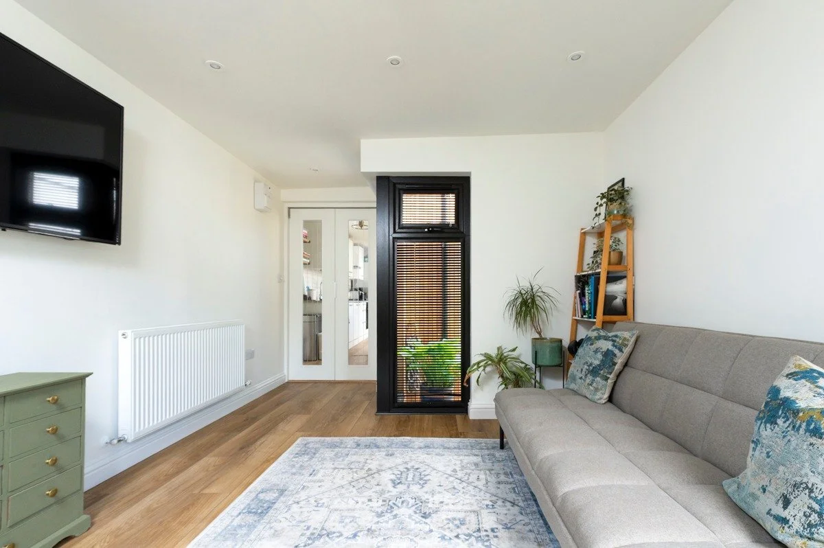 Living room with a gray sofa, potted plants, a wooden shelf with books, a wall-mounted TV, hardwood floors, and a doorway with glass doors leading to another room.