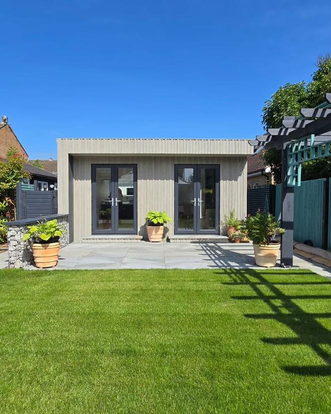 A modern backyard with a small gray building featuring glass double doors, potted plants, a stone patio, a well-maintained green lawn, and a clear blue sky.