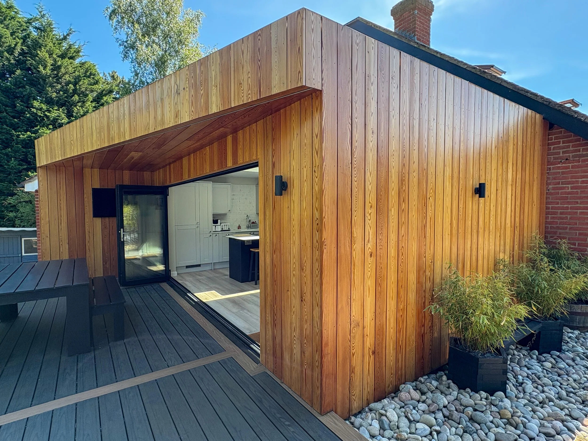 Exterior view of a modern wooden under-rafter extension with a sliding glass door open to reveal a white kitchen inside. The extension is built with vertical wooden panels and features a wooden deck with two black benches, light fixture on the wall, and decorative rocks with plants nearby. The sky is clear and trees are visible in the background.