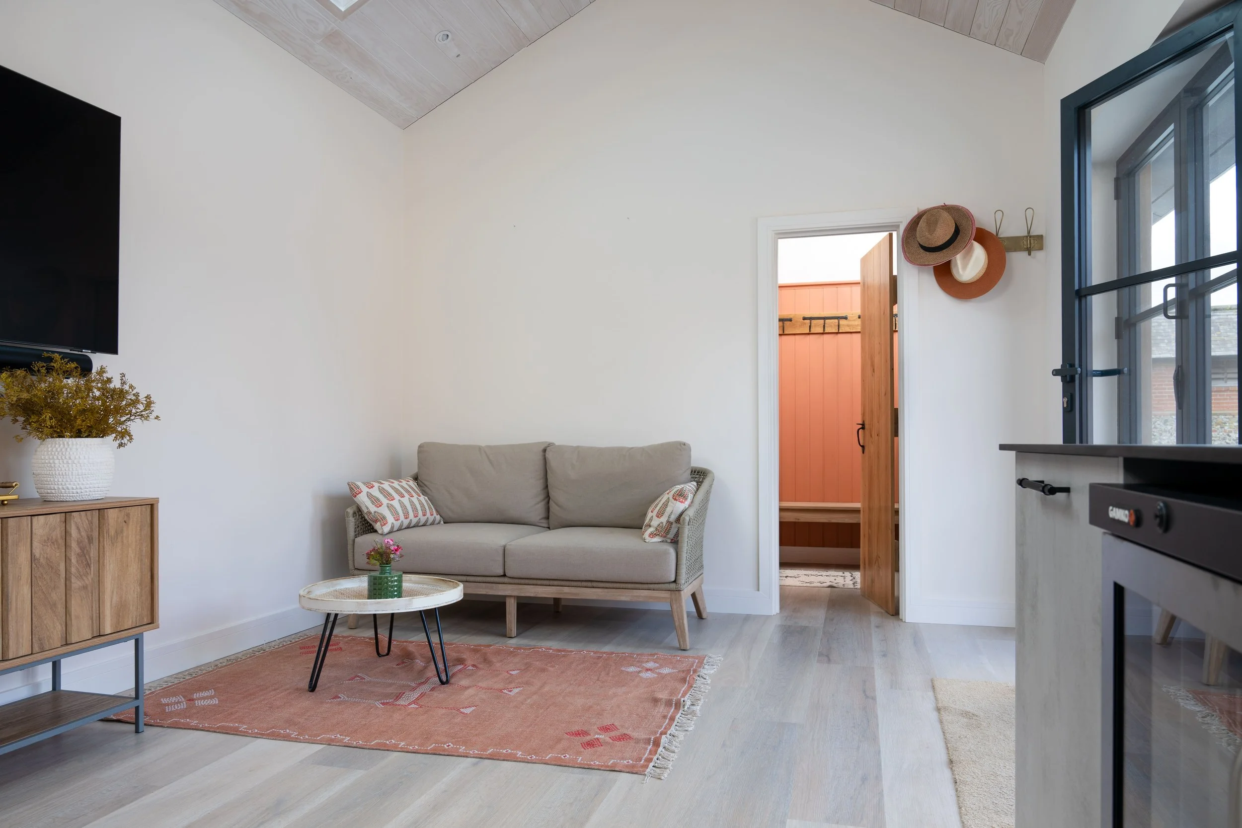 A cozy living room with a beige two-seat sofa, a round coffee table with a small green vase and pink flowers, a pink area rug, a wooden cabinet with a potted plant, wall hooks with hats, a doorway to a small screened-off porch, a window with a blue frame, and a TV mounted on the wall.